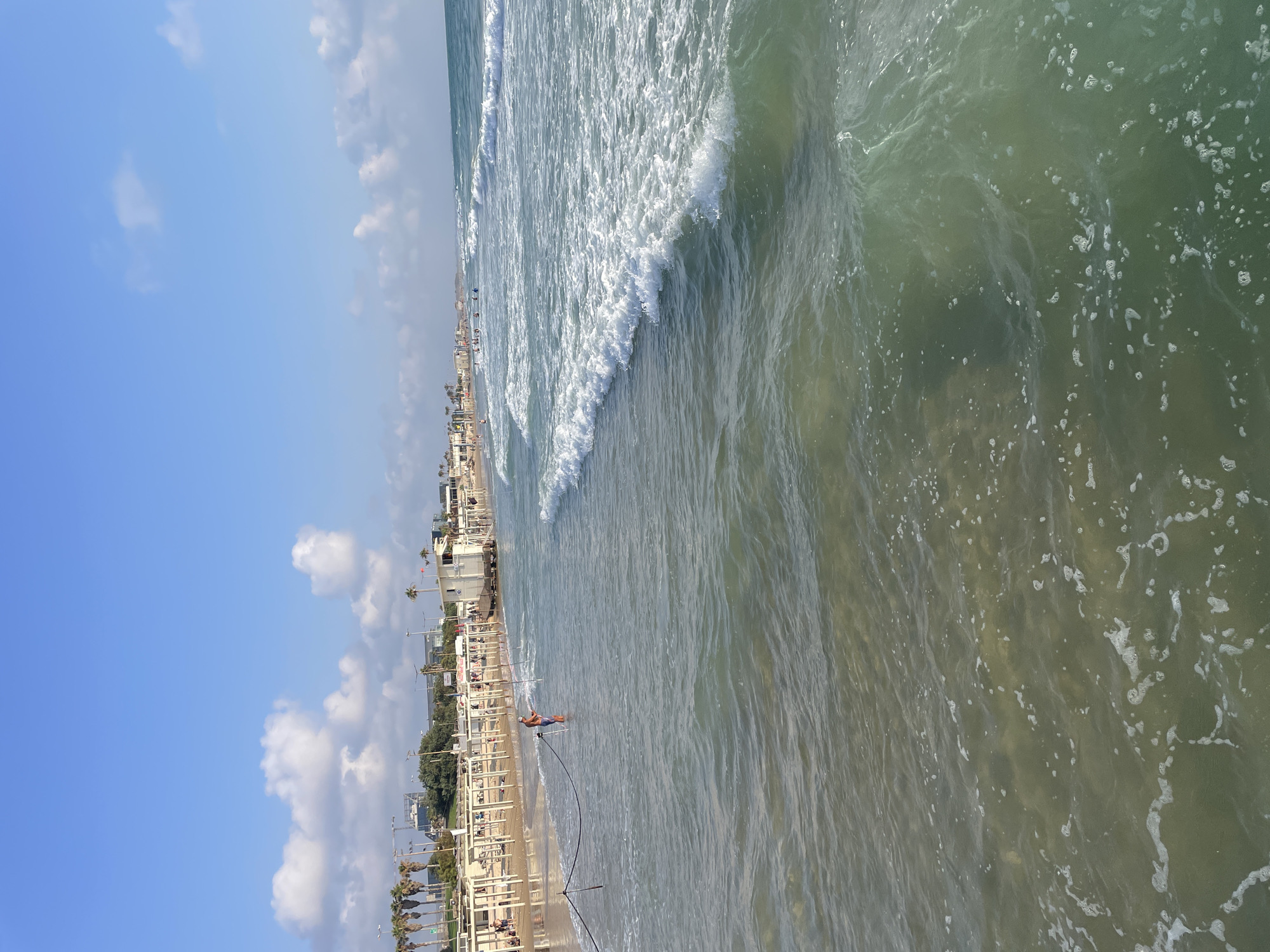The image shows a sunny day at a beach in Haifa, Israel. The foreground is dominated by the ocean, with waves breaking and creating white foam. The water appears clear and greenish-blue. In the mid-ground, there is a sandy beach lined with white cabanas and palm trees. People are visible on the beach and in the water, some swimming and others sunbathing. A lifeguard tower is situated on the beach. In the background, there are buildings and more palm trees, suggesting an urban setting bordering the beach. The sky is blue with scattered white clouds. A person in the water is holding onto a rope that extends to the shore, possibly a fishing line or a safety rope.