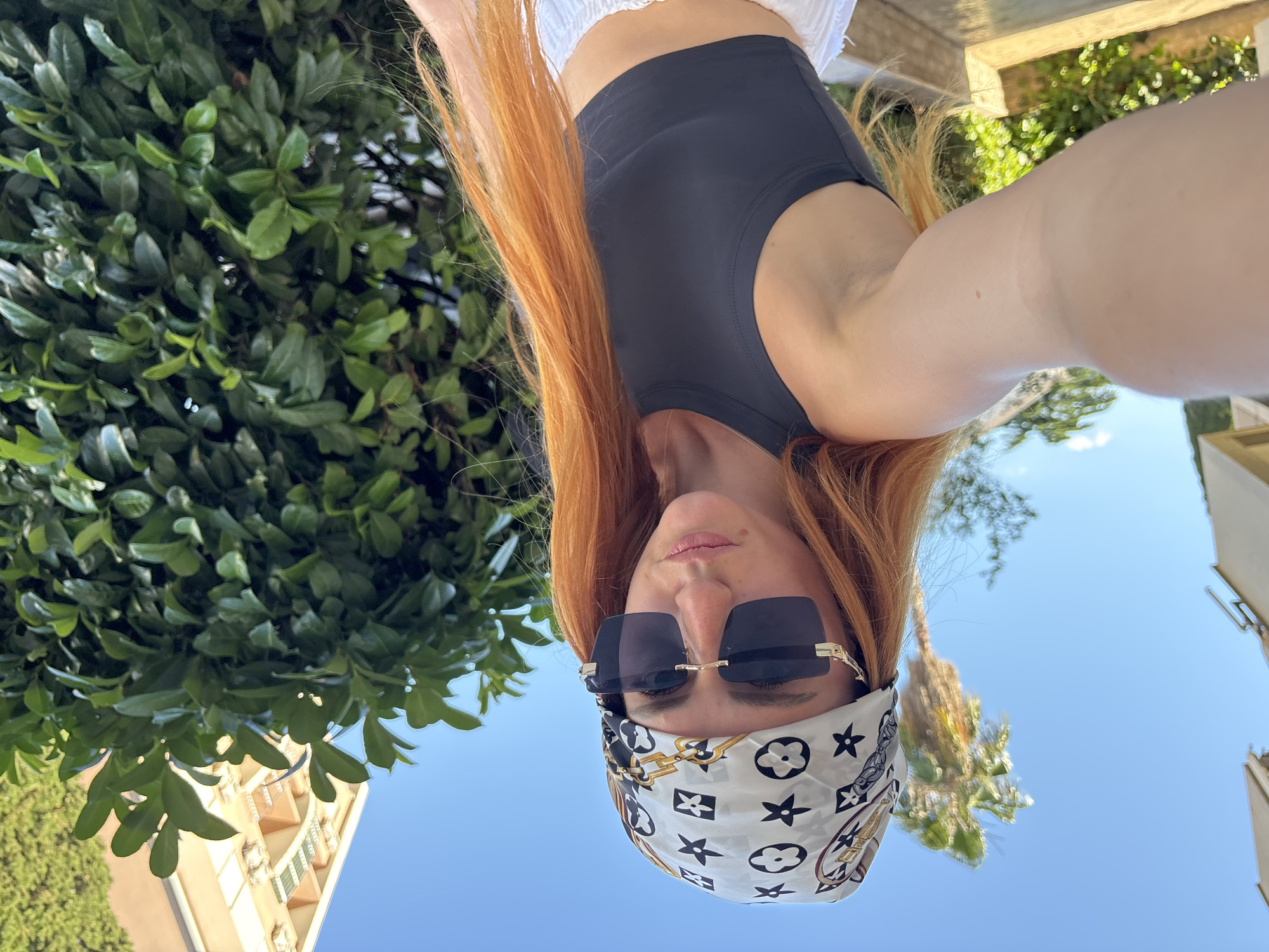 In a medium shot, a woman with long, auburn hair is captured from the shoulders up, facing forward. She wears a black, sleeveless crop top and a white bandana with a black and gold pattern tied around her head. Large, dark sunglasses obscure her eyes, and her expression is neutral. The background features a lush green hedge, a building with balconies, and a palm tree under a clear blue sky, suggesting an outdoor urban setting, likely in Budva, Montenegro, given the architectural style and vegetation. The lighting indicates a sunny day. The image is a selfie, with the woman's left arm extended, holding the camera.
