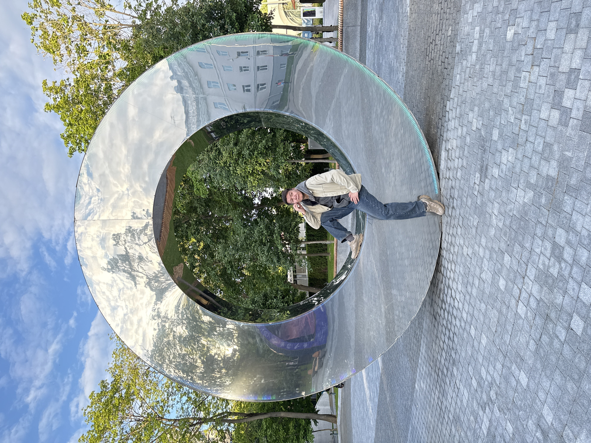 An outdoor scene in Moscow, Russia, depicts a man posing playfully with a large, highly reflective metallic ring sculpture. The man, positioned to the right of the central void of the sculpture, is seen in a dynamic, crouched pose with one knee bent and the other leg extended, looking directly at the camera with a slight smile. He is dressed in a light beige jacket over a dark grey hoodie, blue jeans, and light brown sneakers.
The prominent object is a massive, highly polished, silver-toned ring-shaped art installation. Its mirror-like surface extensively reflects the surroundings. Reflections on the sculpture include a bright blue sky with scattered white clouds on its upper left exterior, while the inside and parts of the exterior showcase lush green trees and foliage. On the upper right exterior, a light-colored building with multiple windows is distinctly reflected. Along the inner edge of the ring, a series of small, multi-colored LED lights (green, blue, and purple) are visible, hinting at modern design.
The ground is paved with light grey, rectangular cobblestones or pavers, extending across the visible area. The scene is illuminated by natural daylight, suggesting late afternoon or early evening with clear weather. The composition captures a moment of playful interaction with urban public art. No visible text is present in the image.
