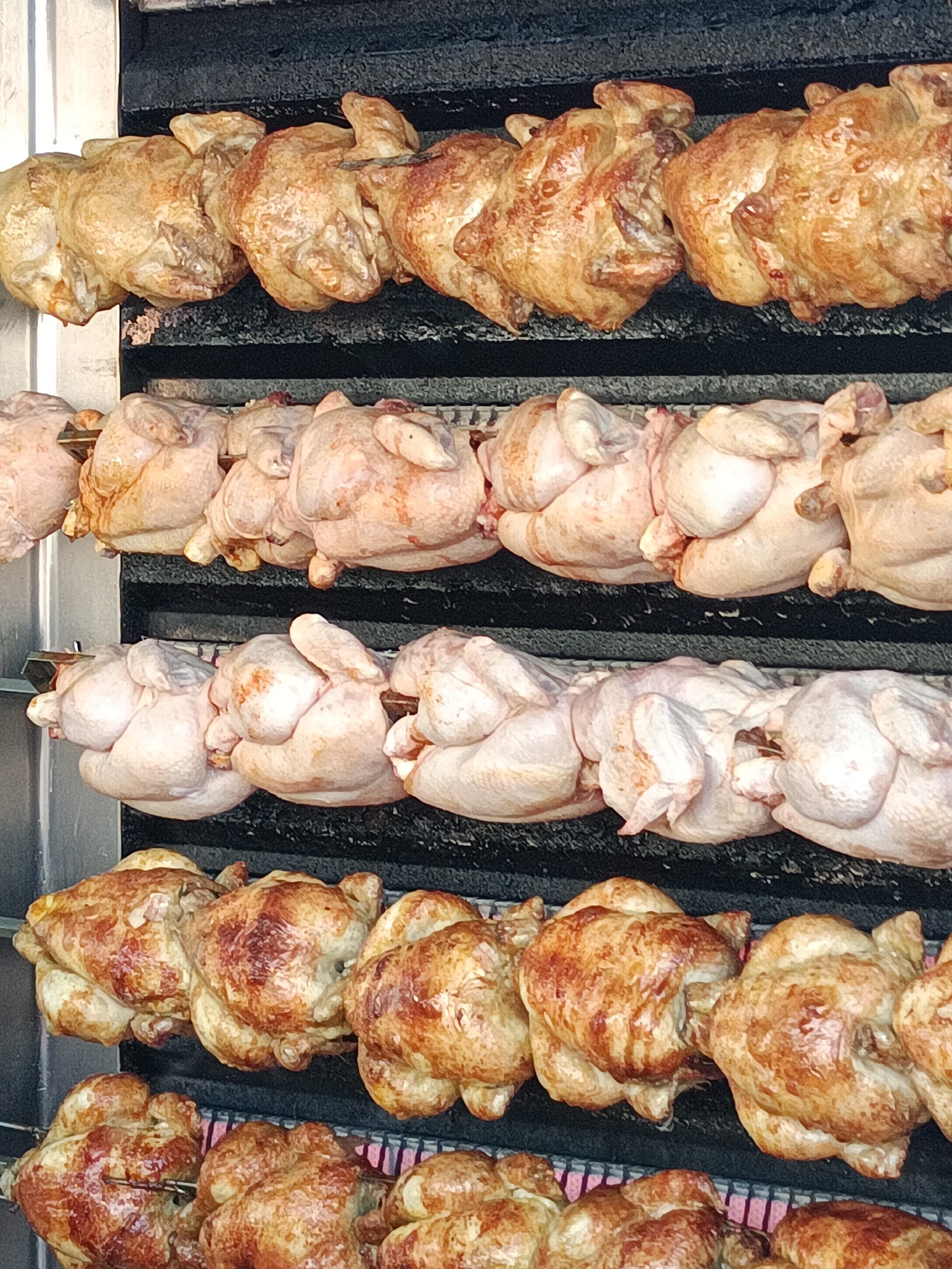 The image shows a rotisserie with multiple rows of whole chickens being cooked. The top two rows feature chickens that are golden brown and appear to be fully cooked, with glistening skin. The chickens are skewered on metal rods that rotate them evenly. Below these, there are two more rows of chickens that are pale pink and appear to be raw or partially cooked. The rotisserie is set against a dark, textured background, likely part of a commercial cooking apparatus. The scene is observed from a slightly elevated, close-up perspective, emphasizing the cooking process. There are no people or animals visible in the image. The location of this rotisserie in Le Neubourg, France, is not specified by any visual cues in the image itself.
