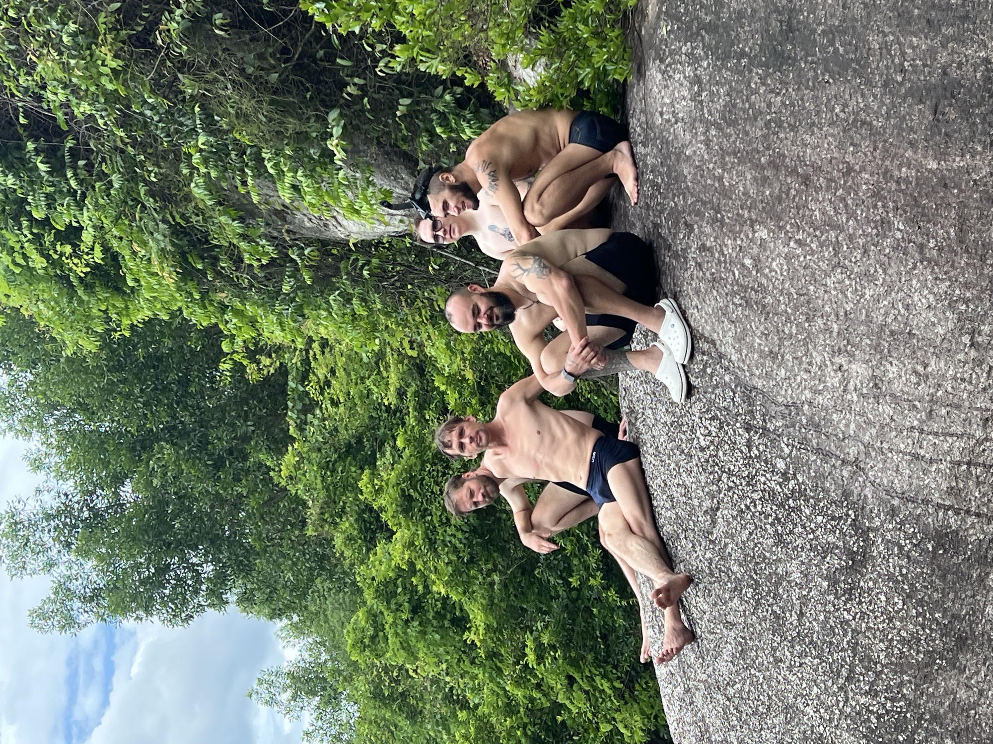 Five adult males are sitting on a large, grey, speckled rock. The rock is situated in a lush, green environment with dense trees and foliage covering a rocky incline behind them. The sky above is partly cloudy with patches of blue.
From left to right:
The first man is lying on his side, propped up on his left elbow, with his legs extended. He is wearing dark blue swim trunks. He has a beard and short brown hair.
The second man is sitting upright, facing forward, with his arms resting on his knees. He is wearing dark blue swim trunks. He has short brown hair and no beard. He appears to be looking towards the camera.
The third man is sitting with his legs crossed, leaning slightly forward. He is wearing dark swim trunks. He has a beard and is bald on top with short hair on the sides. He is wearing a necklace with a pendant. He is looking downwards and smiling.
The fourth man is sitting with his legs tucked in, facing the camera and slightly to the right. He is wearing dark swim trunks. He has dark, short hair and a beard. He is wearing glasses and a snorkel mask pushed up onto his forehead. He is smiling.
The fifth man is sitting with his legs extended, facing the camera and slightly to the right. He is wearing dark swim trunks. He has short dark hair and a beard. He is wearing white Crocs-style shoes. He is looking towards the camera and smiling.
The location is identified as being in the Marble Mountains (Ngũ Hành Sơn) in the city of Đà Nẵng, Vietnam. The distinctive rock formations and abundant vegetation are characteristic of this area.