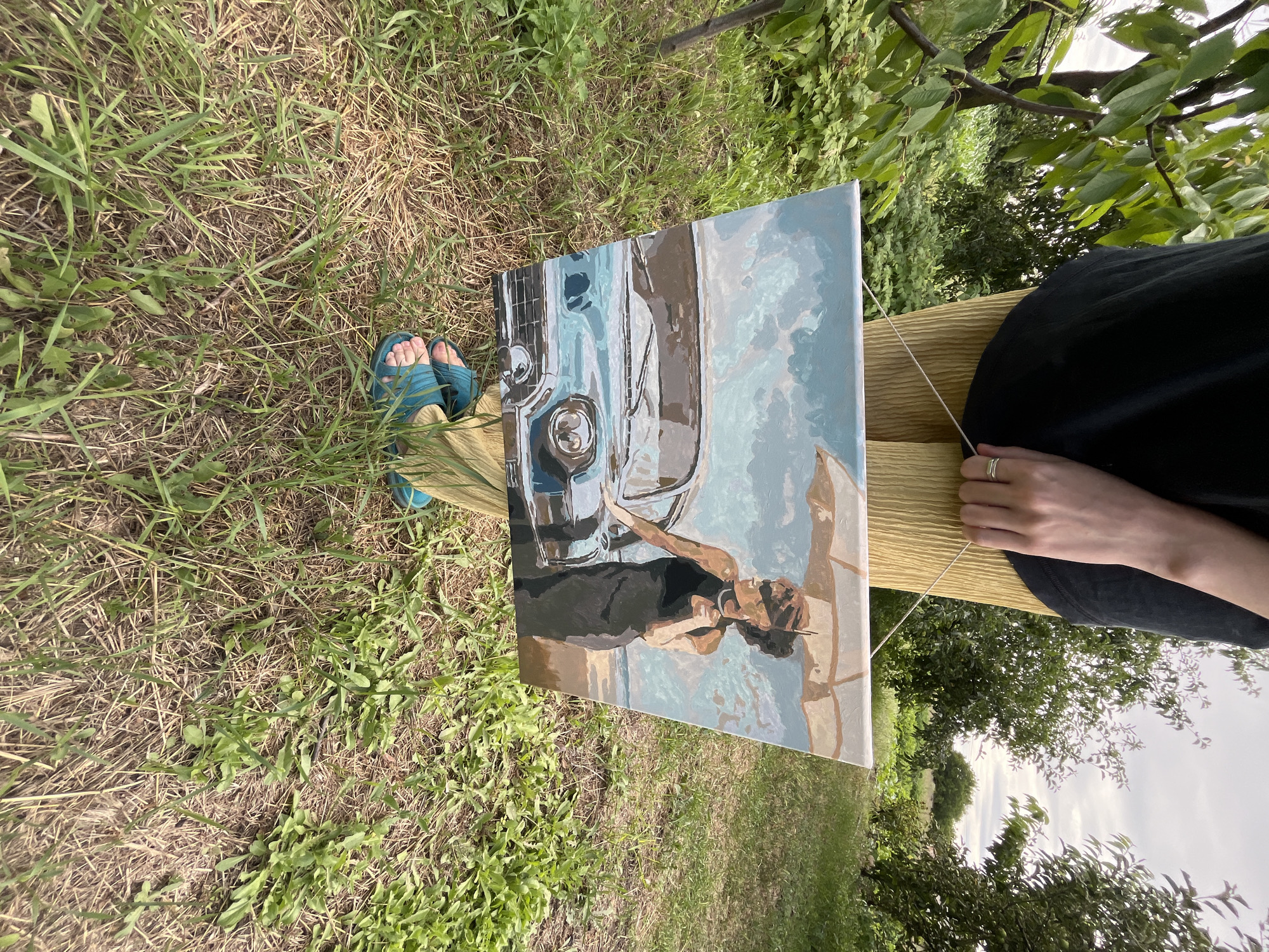 A person is sitting outdoors, possibly in a garden or park, holding a painting of a vintage blue car and a woman with an umbrella. The painting is tilted, allowing a partial view of its contents. The person's feet, clad in blue sandals, are visible near the bottom left of the image, resting on dry grass. A thin string or wire connects the painting to a wooden surface, possibly a bench or table. The person's hands, adorned with a ring on one finger, are also visible, holding the string. The background consists of green foliage and trees, suggesting an outdoor setting during the day, with a cloudy sky overhead. The scene evokes a sense of creativity and relaxation, with the person engaged in an artistic endeavor amidst nature.