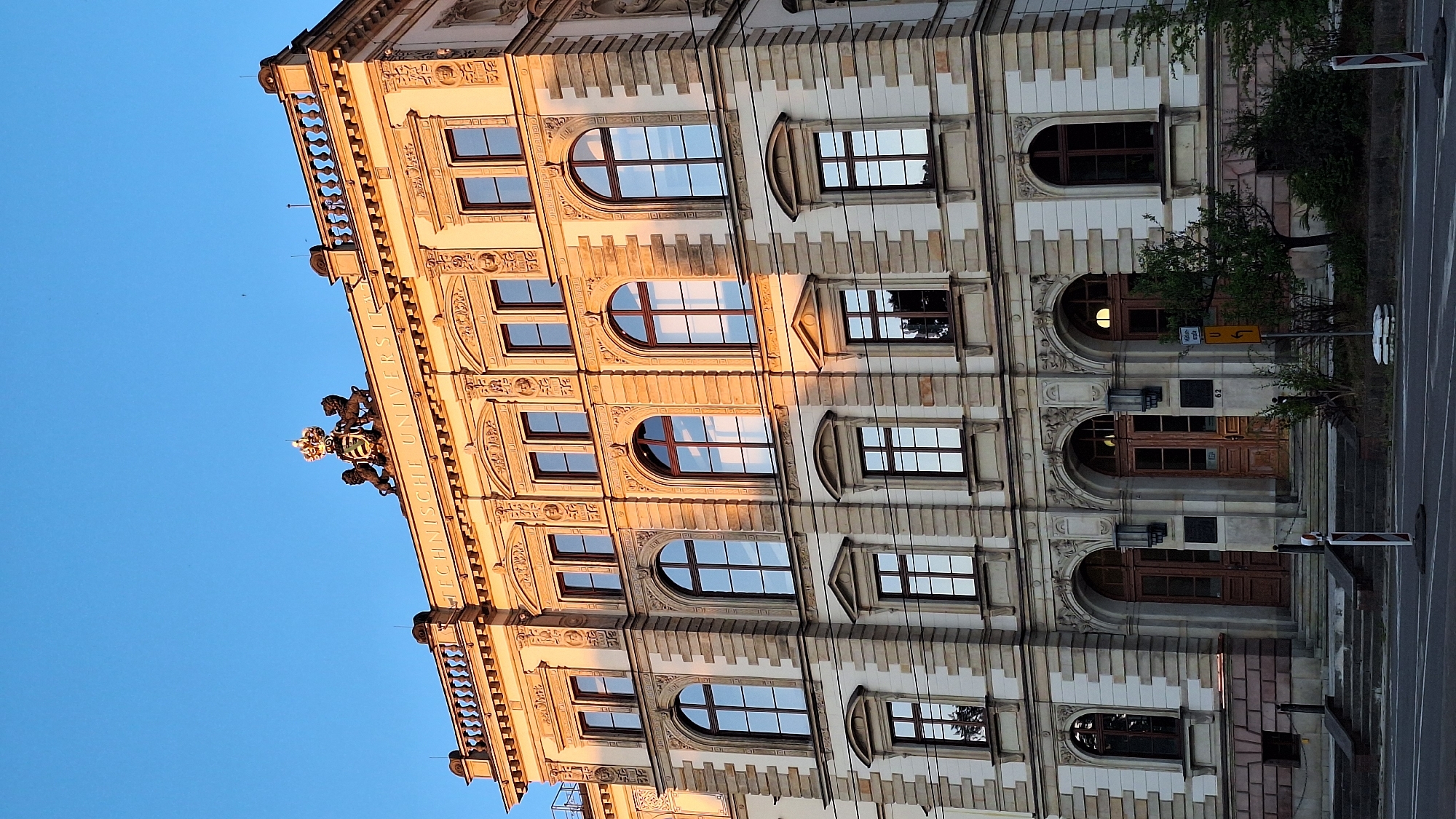 This is a low-angle, sideways shot of a grand, classical building with an ornate facade, illuminated by the warm glow of what appears to be late afternoon or early evening sunlight. The building has multiple stories, characterized by a series of arched and rectangular windows with dark wooden frames. Many windows reflect the clear blue sky and the surrounding architecture, creating a sense of depth and continuity. The facade is detailed with stone carvings, decorative moldings, and recessed panels. At the top of the building, a balcony or balustrade runs along the edge, and above it, a crest with lions and a crown is visible. The text "TECHNISCHE UNIVERSIT" is inscribed on the building's facade.
The scene includes elements of a street or pathway in front of the building, with trees and foliage growing near the entrance. A yellow traffic sign with an arrow is present, and the number "62" is visible above one of the large wooden doors. The overall impression is one of historical significance and architectural beauty, bathed in the gentle light of a fading day. The clear sky suggests fair weather. No people are visible in the frame.