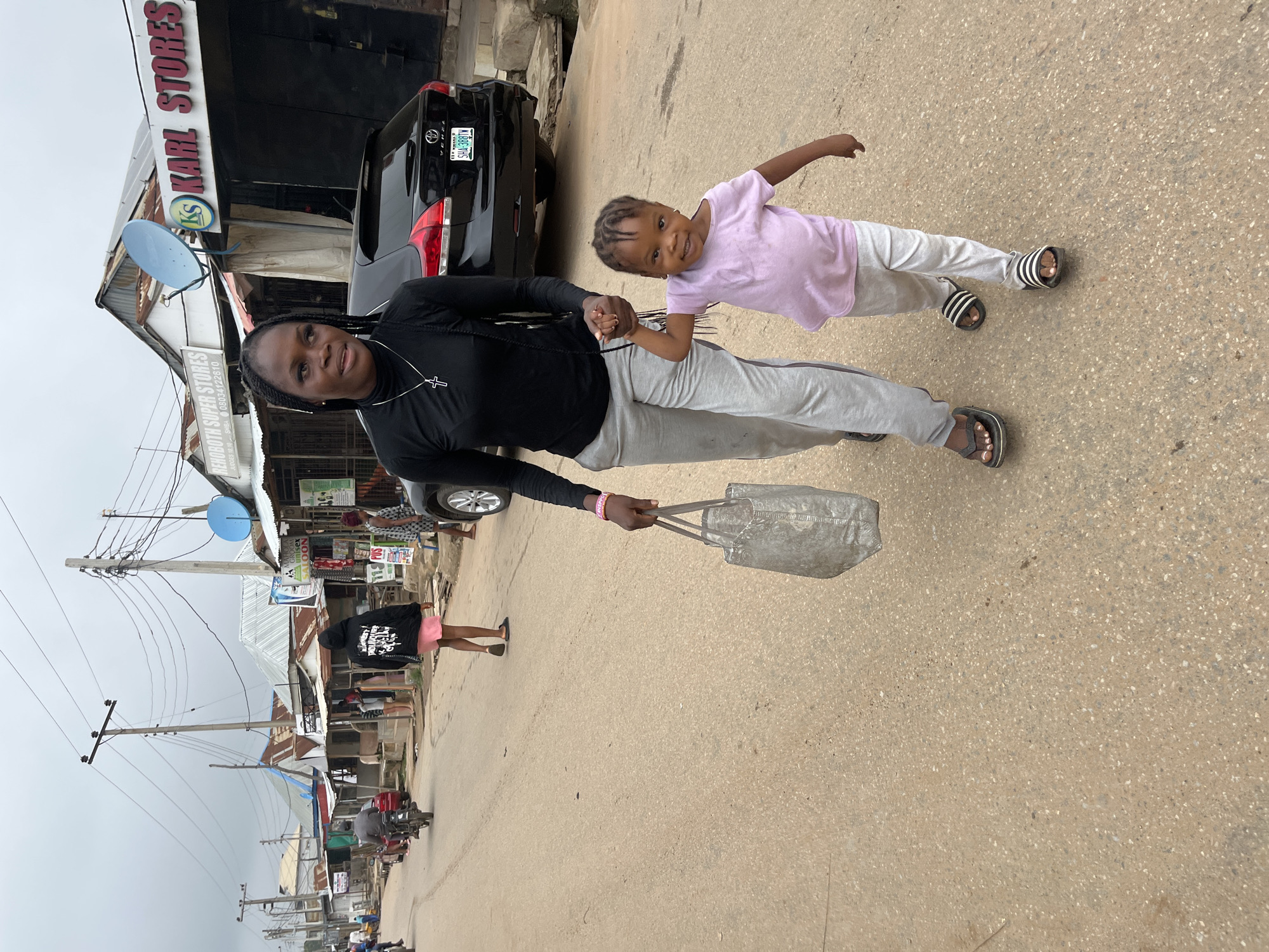 A Nigerian woman and a young child walk hand-in-hand down a dirt road in Abuja, Nigeria. The woman is dressed in a black long-sleeved shirt and grey sweatpants. She carries a large grey tote bag in her left hand. The child, wearing a light pink shirt and grey sweatpants, smiles and looks towards the camera. In the background, a black SUV is parked, and various market stalls and shops line the street, including "Karl Stores" and "Renoboth Super Stores." The sky is overcast, and utility poles with numerous wires are visible. Other people and a motorcycle can be seen further down the road.