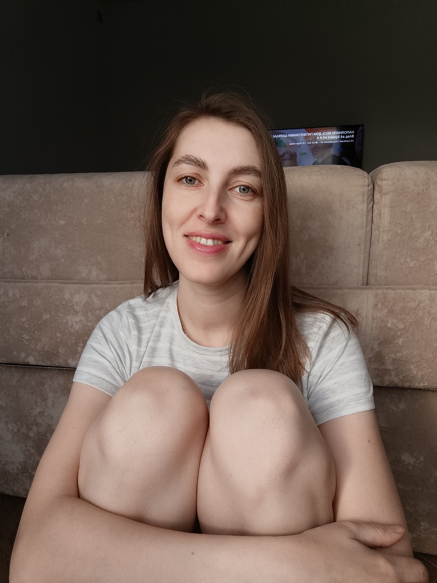 The image features a young woman seated indoors, likely in a home in Chaikovsky, Russia. She is positioned on the floor or a low surface, facing the camera with a pleasant, relaxed smile. Her long, light brown hair falls around her shoulders, and her eyes are light green or blue. She wears a light grey t-shirt with subtle horizontal stripes. Her knees are drawn up towards her chest, and her arms are wrapped around them.
Behind the woman is a light-colored, upholstered sofa or couch, serving as the backdrop. The room appears dimly lit, typical of an indoor setting during the day without direct bright sunlight. In the upper right background, a television screen is visible. The screen displays mirrored and largely illegible text, which appears to be Cyrillic script, along with some graphics and what could be a timeline or progress bar. The specific content playing on the television is not discernible. The overall scene suggests a casual and comfortable atmosphere, with the woman conveying a friendly and approachable demeanor.