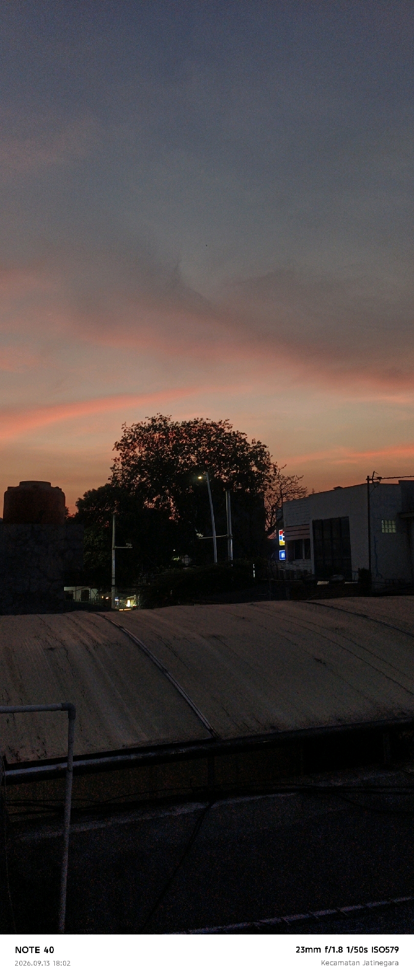 A photograph taken at dusk shows a silhouetted tree and buildings against a sky filled with pink and grey clouds. A water tower is visible on the left. In the foreground, a dark, textured rooftop dominates the lower portion of the image. Text overlays indicate the location as Kecamatan Jatinegara in East Jakarta, Indonesia, and the camera settings as 23mm f/1.8 1/50s ISO579. The timestamp reads 2026.09.13 18:02.