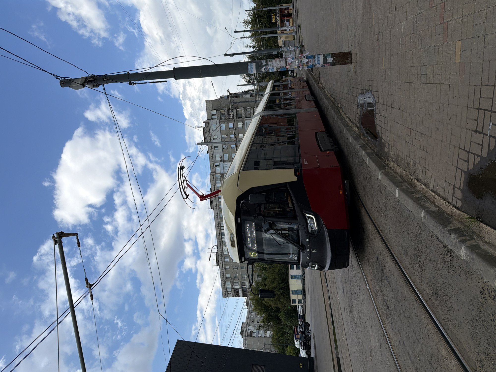 A modern, red and cream tram with headlights on is moving along tram tracks on a city street. The tram has the number 2853 visible on its front. The tram is on the right side of the frame, with the tracks extending into the distance. The street has a paved sidewalk with a curb and a few puddles on the ground, indicating recent rain. In the background, there are multi-story residential buildings, trees, and utility poles with overhead wires. The sky is partly cloudy with blue sky visible. The scene appears to be in an urban environment, possibly a residential or commercial district. Based on the text on the tram and in the image, this is likely a tram in Nizhny Novgorod, Russia.