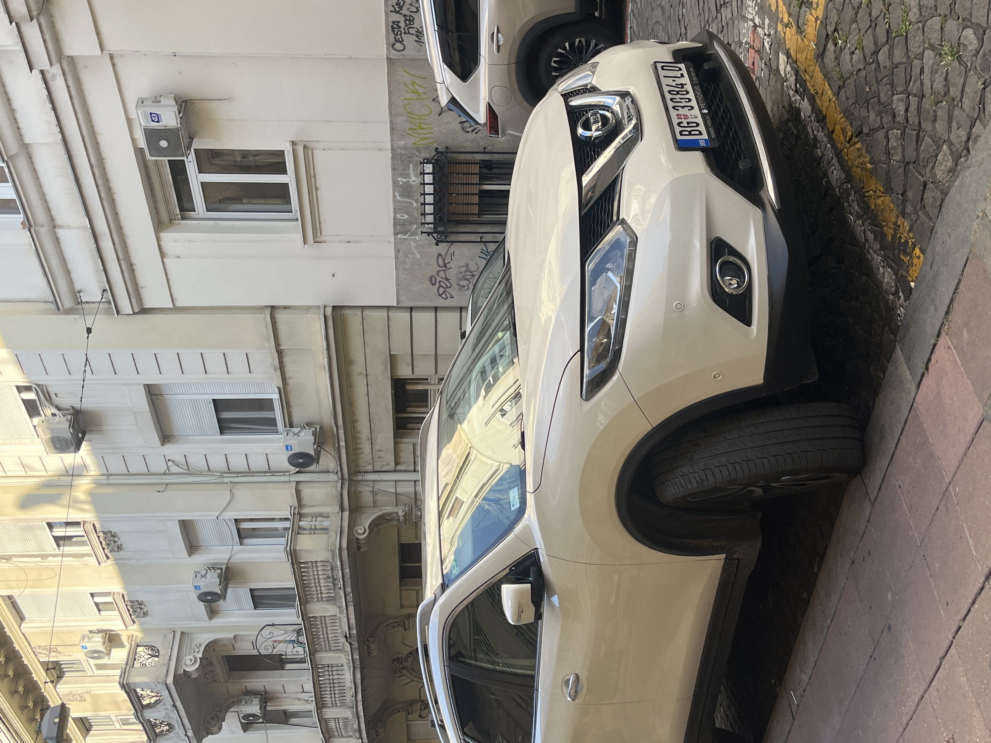 The image captures a street scene in Belgrade, Serbia, focusing on a beige Nissan Qashqai parked on a cobblestone street. The car is positioned in the foreground, partially obscuring a second beige SUV parked behind it. The building facade in the background is light-colored, featuring multiple windows with shutters and several air conditioning units mounted on the exterior walls. Graffiti is visible on the lower section of the building. The street itself is paved with cobblestones and marked with yellow lines indicating parking restrictions. The lighting suggests it is daytime, with sunlight casting shadows on the building. No people are visible in the frame.