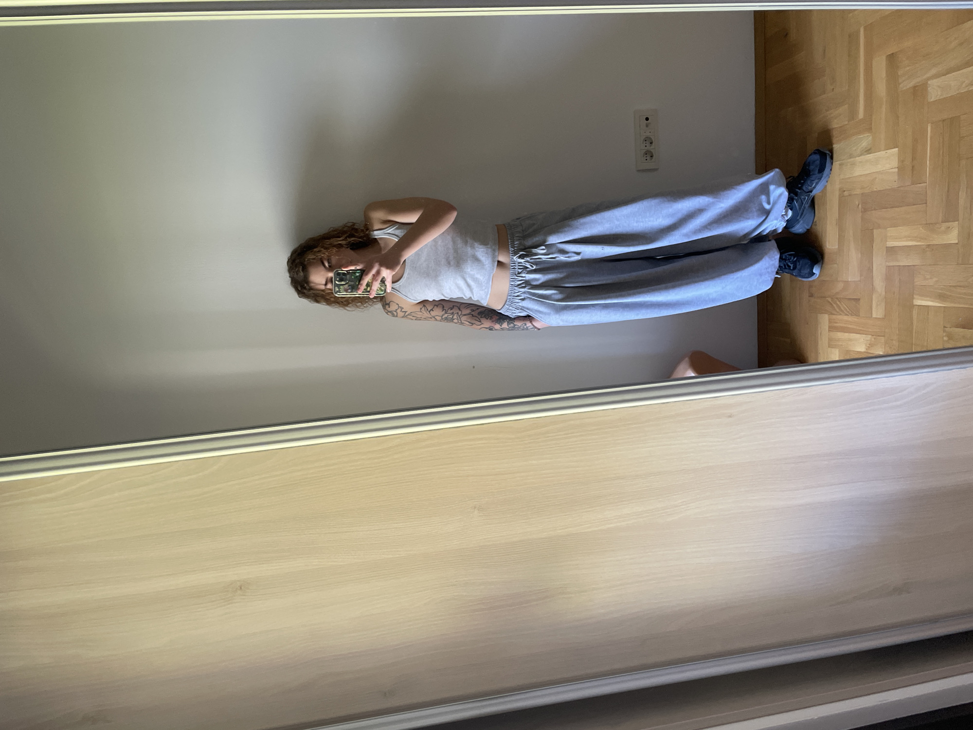 The image is a mirror selfie of a person with curly brown hair, wearing a gray tank top and baggy gray sweatpants, and dark sneakers. The person has tattoos on their left arm and is holding a smartphone in their left hand. The setting appears to be an interior space with light-colored wooden floors and light gray walls. There's an electrical outlet visible on the wall. The mirror frame is made of light wood with a silver trim. The location, Boreti, Montenegro is not identifiable from the image.