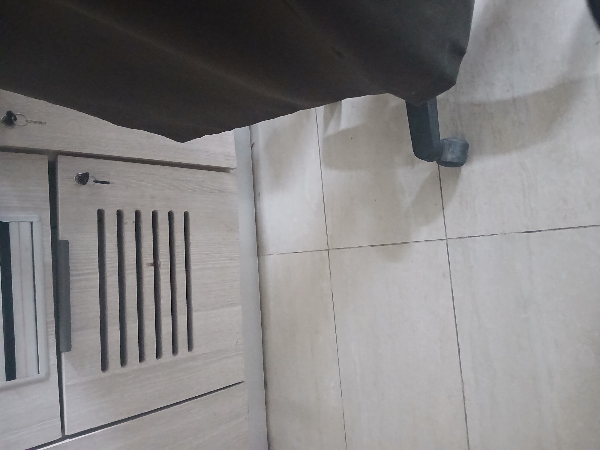The image shows a close-up view of a wooden cabinet with vents, a key in a lock, and a dark fabric object, likely part of a chair, in the foreground. The cabinet is light-colored with a visible wood grain. A horizontal vent with multiple slats is prominent on the lower section of the cabinet. A small key with a keychain is inserted into a lock on the right side of the cabinet. To the right of the cabinet, a large expanse of dark fabric obscures much of the scene. The floor is tiled with large, light-colored square tiles. The overall impression is of an indoor environment, possibly an office or storage area. No specific location in Peshawar City Tehsil, Pakistan, can be identified from the visual information provided.