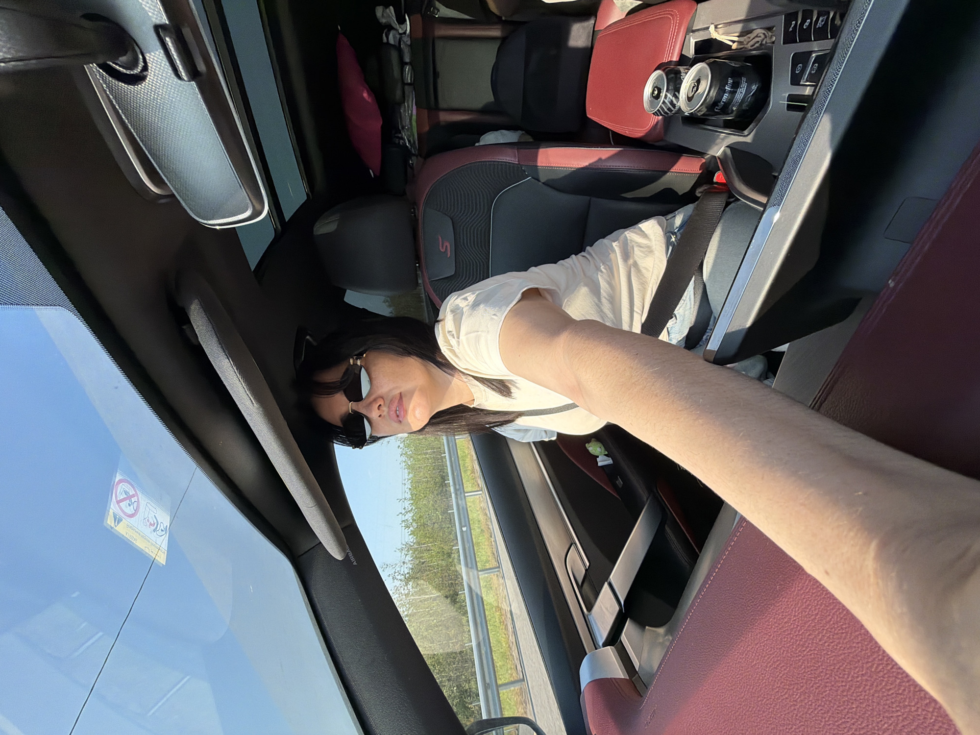 The image shows the interior of a car. A woman wearing sunglasses and a white t-shirt is in the driver's seat. She has dark hair and is looking towards the right. The car has red and black leather seats, and there are two cans of soda in a cup holder. The car is on a road with trees and a guardrail visible outside the window. The sky is clear and blue. The image was taken in Fyodorovo, Russia.