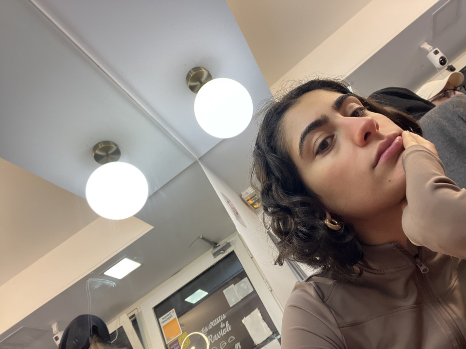A woman with curly dark hair and gold hoop earrings rests her cheek on her hand. She is wearing a brown long-sleeved top. The background shows a ceiling with two round lights and a door with a sign that says "Ravioli." A security camera is visible in the corner. Other people can be seen blurred in the background.