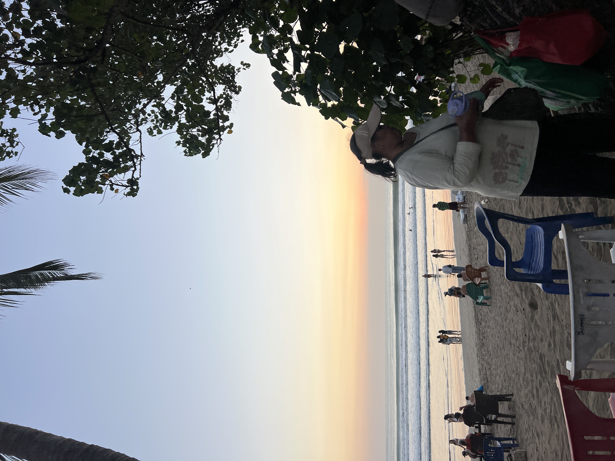 The image captures a serene beach scene in Kuta, Indonesia, during what appears to be sunset. The sky transitions from a soft blue at the top to hues of yellow and orange near the horizon, where it meets the calm ocean with gentle waves. Several individuals are present on the beach, some seated on chairs, while others are walking or standing by the water's edge. In the foreground, a woman wearing a visor and a light-colored shirt is visible, holding a small blue object. She is standing next to a cluster of green and red shopping bags. The beach itself is sandy, with a few palm trees and lush foliage providing a natural frame to the scene. The overall atmosphere is peaceful and relaxed, indicative of a typical evening on a popular Indonesian beach.