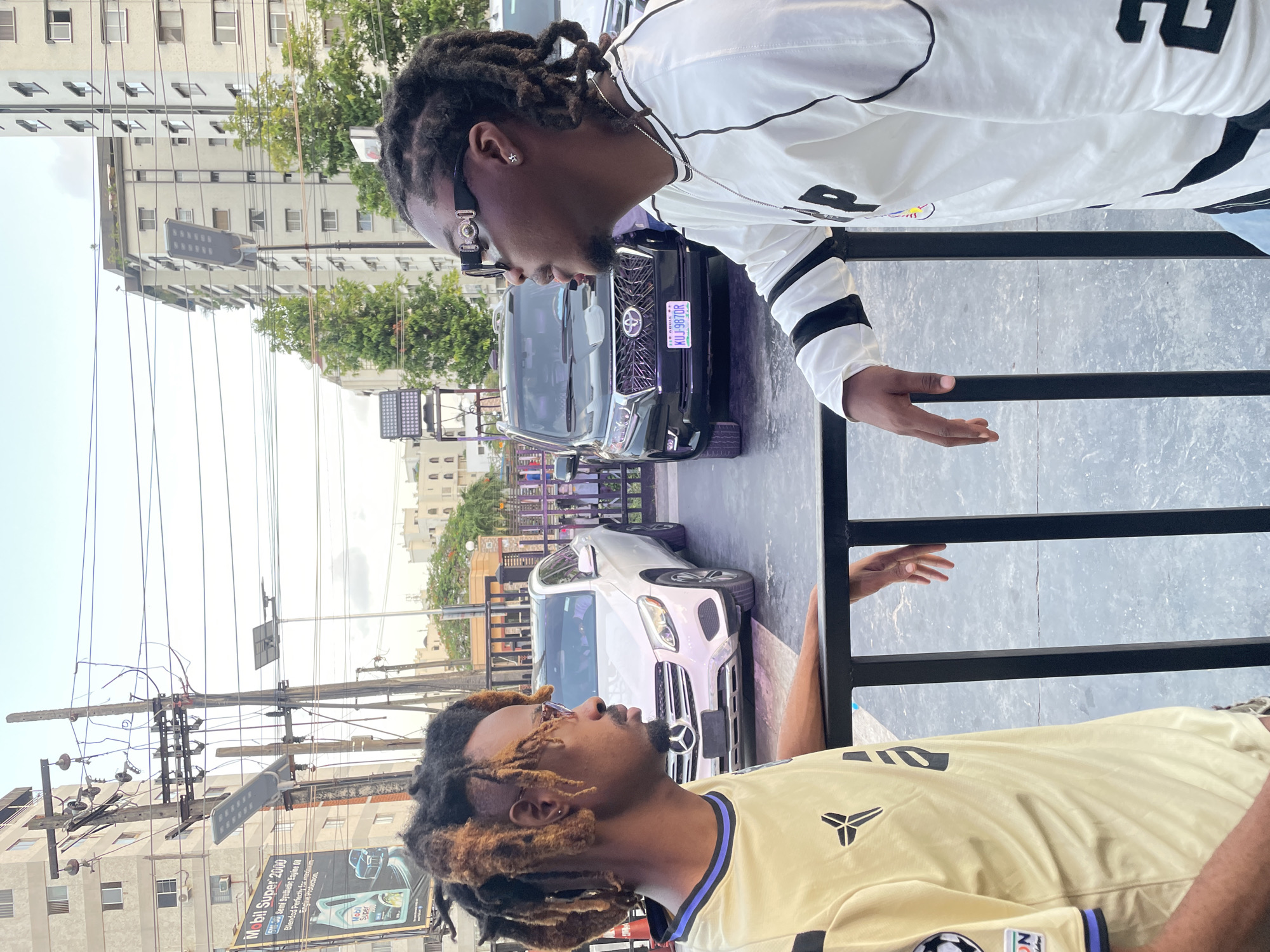 Two men are talking on a patio behind a black metal railing. One man is wearing a light yellow shirt, and the other man is wearing a white long-sleeve shirt. The city has light-colored buildings and is densely populated. Two cars are parked, one silver and one black, and the black car's license plate says "KUJ 987 DR" with a Nigerian flag. There is a sign for "Mobil Super 2000" that says "Semi Synthetic Engine Oil, Blended Perfectly for maxi Engine Protection." The city and state are unknown.