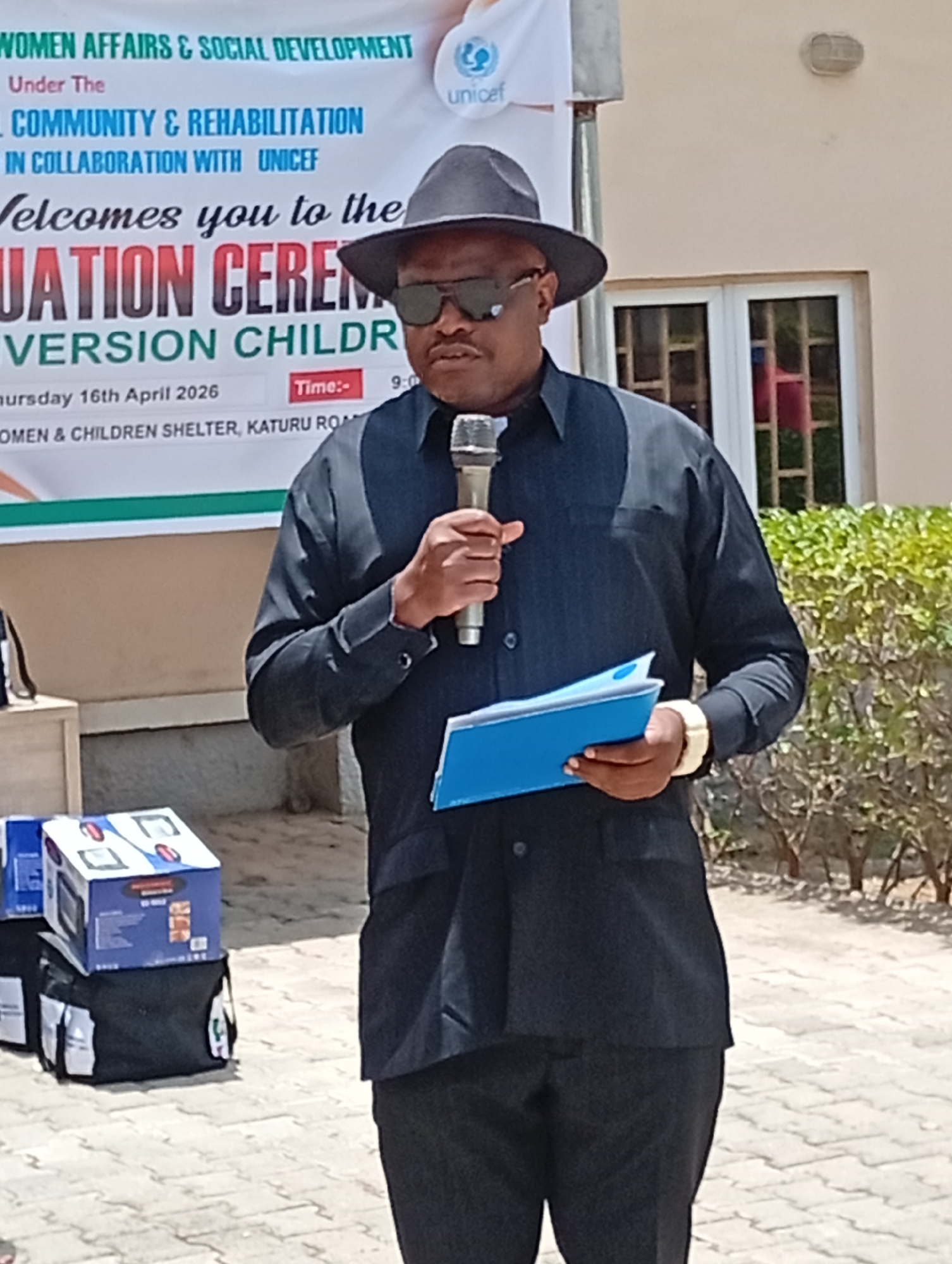 The image depicts an outdoor event taking place in Unguwar Sarki, Nigeria, under bright daylight conditions. A man stands centrally, speaking into a microphone held in his right hand. He is dressed in a dark long-sleeved shirt and trousers, complemented by a dark fedora-style hat and sunglasses. In his left hand, he holds a blue folder or binder. A prominent gold-colored wrist watch is visible on his right wrist.
Behind the speaker, a white banner serves as the backdrop, displaying event details. The banner indicates the event is organized by "WOMEN AFFAIRS & SOCIAL DEVELOPMENT" and focuses on "COMMUNITY & REHABILITATION" in "COLLABORATION WITH UNICEF," whose logo is also visible. Text on the banner reads, "Welcomes you to the GRADUATION CEREMONY" relating to "DIVERSION CHILDRE(N)." The specified date for the event is "Thursday 16th April 2026," with a "Time:- 9:0(0)" AM. The location is identified as "WOMEN & CHILDREN SHELTER, KATURU ROAD."
To the left of the speaker, on the paved ground, are several blue and white boxes, possibly containing electronic equipment, stacked on top of black bags. These items may be for display or distribution at the ceremony. The immediate surroundings include a paved ground surface and part of a light-colored building with windows in the background, along with some distant green foliage.