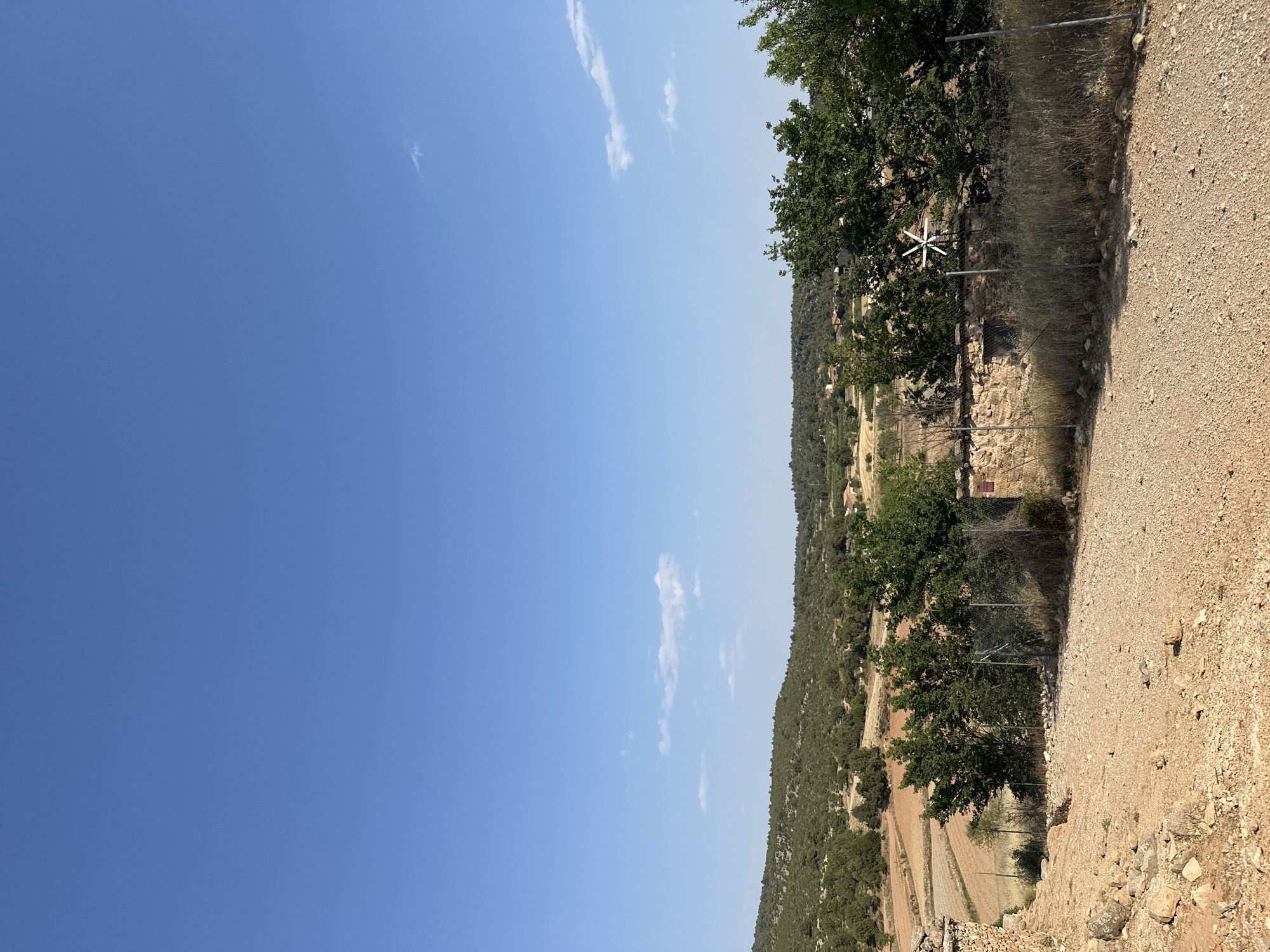 The image shows a rural landscape with a clear blue sky and scattered white clouds. The foreground is a dusty path with gravel and rocks. To the left, there is a stone wall. Beyond the path, there is a fenced area with dry grass and a few trees. In the background, there are rolling hills covered with green vegetation and some cultivated fields. In the distance, small buildings with red roofs are visible. A small white windmill is also visible in the middle ground. This scene is located in the municipality of Alpuente, Spain. There are no people or animals visible in the image.