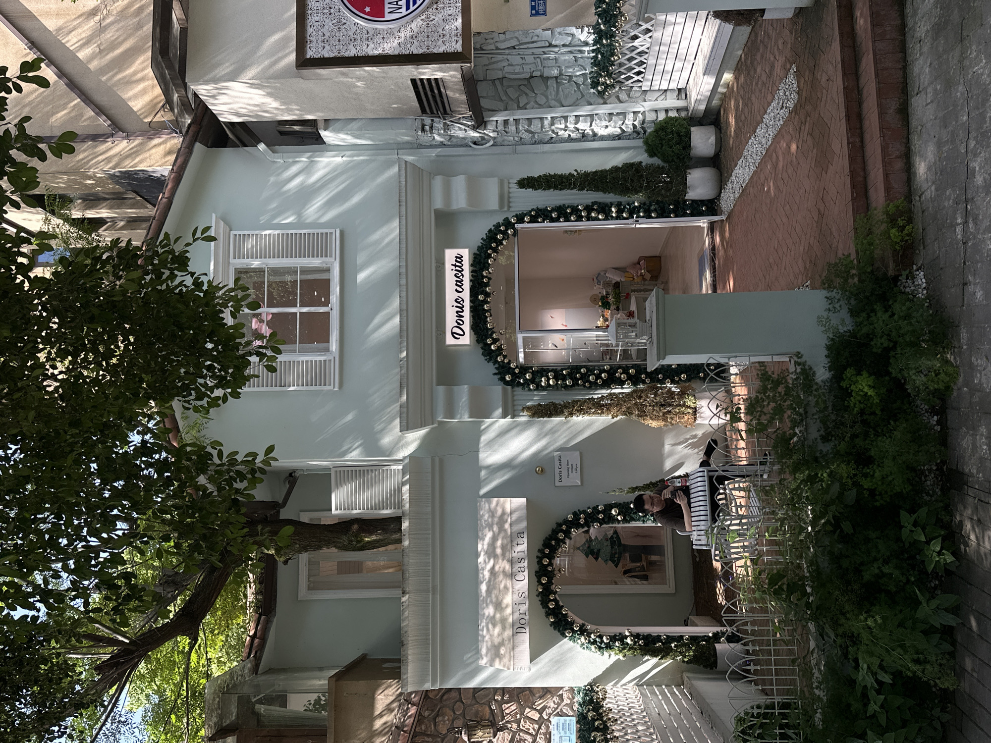 The image depicts the exterior of a store called "Doris Casita" in Liangjiang, China. The store has a pale blue facade with white window frames and shutters. The entrance is decorated with a Christmas-themed garland with gold ornaments. There is a sign with the store's name above the entrance. To the right of the entrance, there is a painting with a similar Christmas garland.
A man is sitting on a chair in the garden in front of the store. The area in front of the store is decorated with white gravel, brick pavers, and potted plants. A large tree is partially visible on the left side of the image.