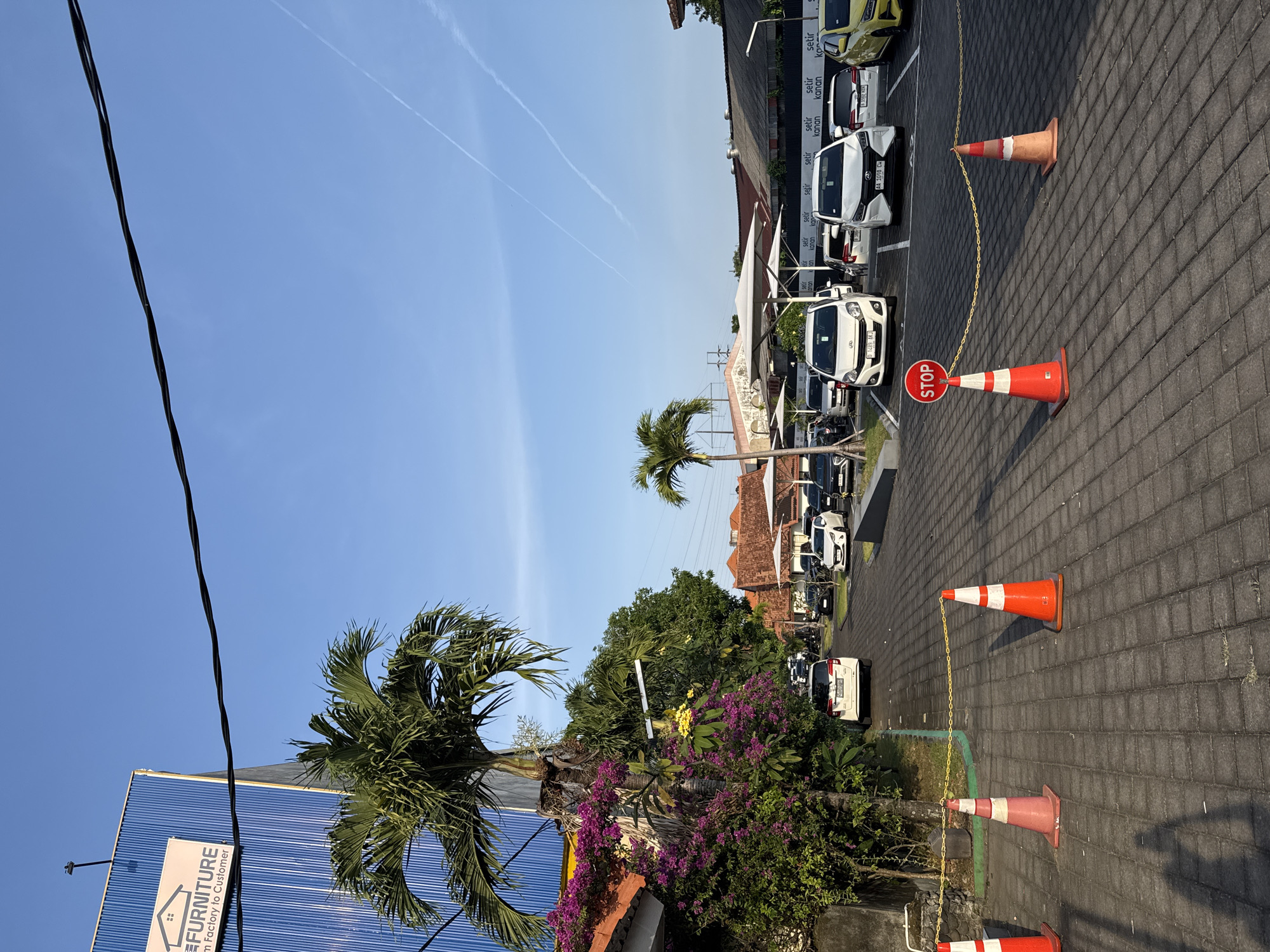 In Denpasar, Indonesia, a parking lot is depicted under a clear blue sky. The lot is paved with interlocking gray bricks. Several white cars are parked in designated spots. Traffic cones, some orange and white, and one with a red "STOP" sign, are placed along the edge of the parking area, marking a boundary. A yellow chain is strung between some of the cones. In the background, buildings with red tile roofs are visible, along with some trees, including a prominent palm tree. A large blue building with a white sign that reads "FURNITURE" is on the left side of the image. The scene appears to be an outdoor commercial area, likely a furniture store's parking lot. No people are visible in the image.