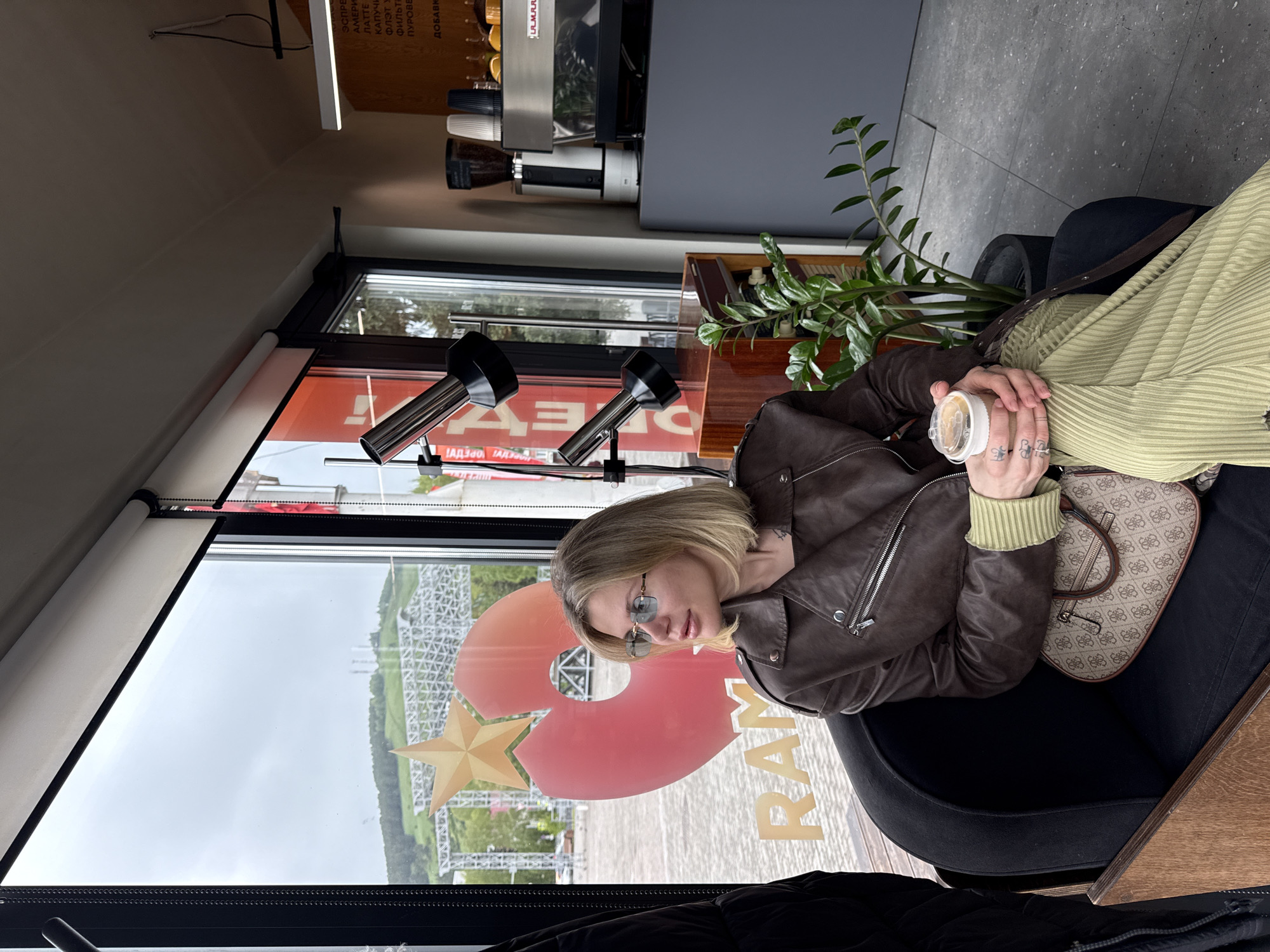A woman with short blonde hair, wearing sunglasses and a brown leather jacket, is seated indoors holding a cup. She also has a green sweater. She is sitting in a black armchair and a patterned handbag is sitting next to her. Outside the window, a large red and gold logo is visible, likely "RAM," along with a glimpse of a roller coaster structure. A light fixture and a coffee machine are visible inside the room, along with a tall green plant. The text displayed on the window includes the word "RAM," suggesting this is the location "Rams Park" of the city of Gorno-Altaysk, Russia.