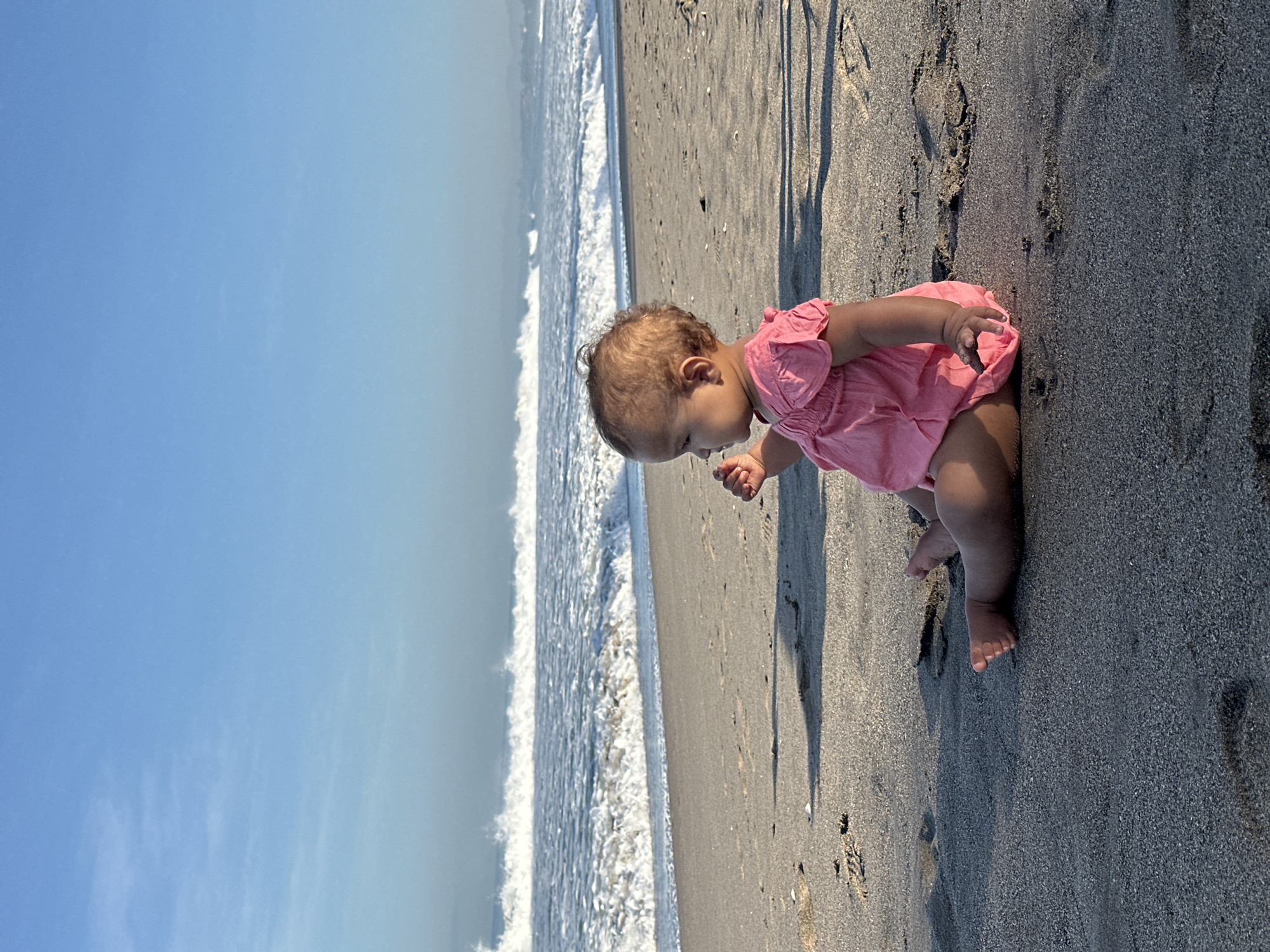 A toddler in a pink dress sits on a sandy beach facing right, looking out at the ocean. The ocean has white waves crashing onto the shore. The sky is a clear blue with a few wispy clouds. The sand is dark and wet near the water, and drier further up the beach. The image appears to be taken during the daytime. The location is not specified within the image itself.