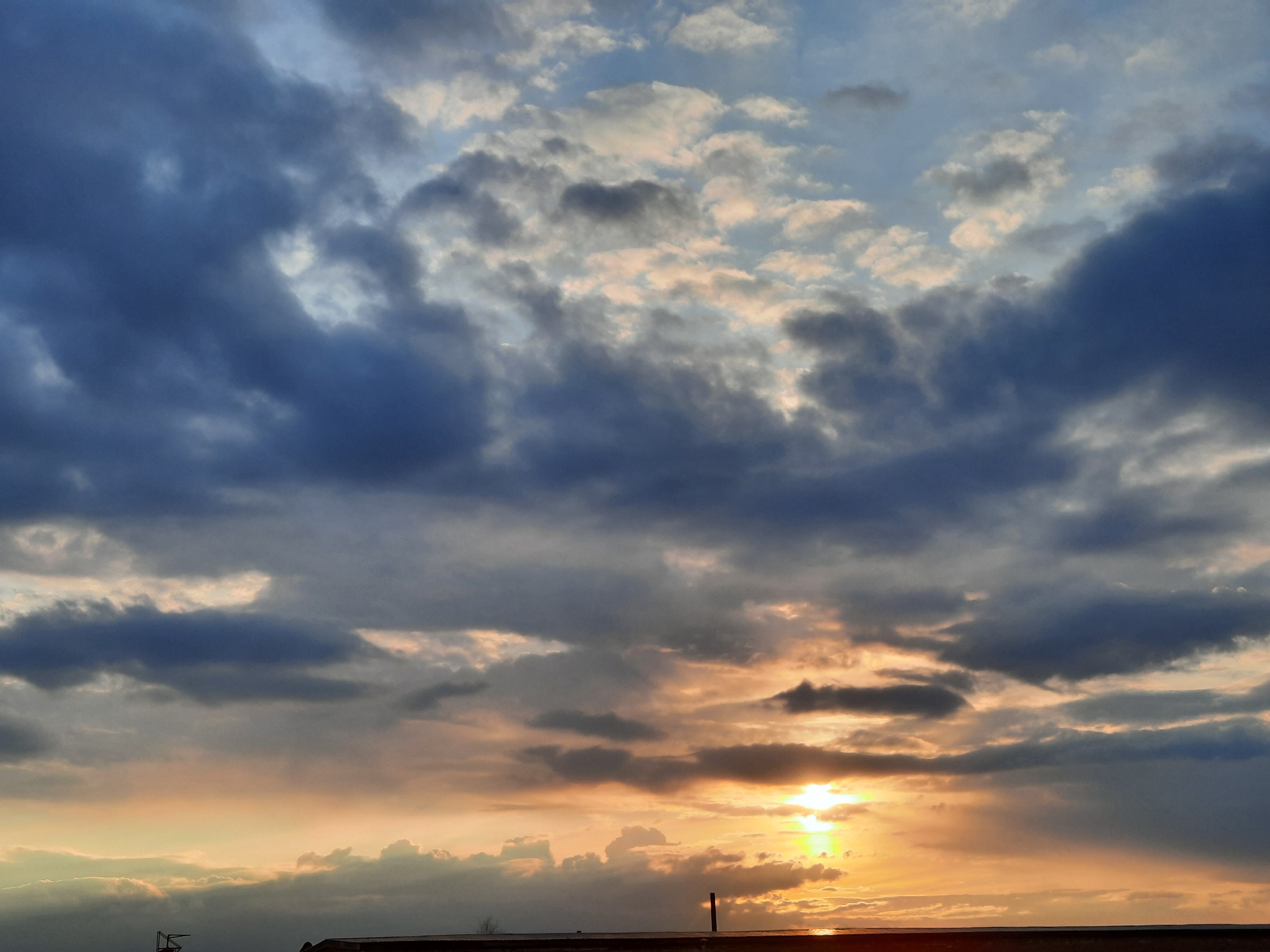 The image displays a vast expanse of sky at either sunrise or sunset over Krasnogorskoye, Russia. The scene is dominated by numerous cloud formations, creating a striking visual contrast between light and shadow.
In the upper and left portions of the frame, dense, dark clouds in shades of blue and deep gray are prominent, suggesting a heavy cloud cover. Towards the middle and lower right, the sun is visible as a brilliant, golden-yellow light source, partially veiled by these clouds. This intense illumination casts warm hues of orange, yellow, and soft pink onto the undersides of the clouds closer to the horizon, creating a vibrant glow that diffuses across the sky.
A narrow, dark line at the very bottom of the image represents a flat horizon, likely indicating a distant landmass or body of water typical of a rural setting. Faint, indistinct dark silhouettes, possibly distant structures or sparse vegetation, are barely discernible against the bright glow of the sun.
No people are visible in the image, nor are there any prominent man-made objects other than the potential faint silhouettes at the horizon. No text is visible. The time of day is clearly twilight (sunrise or sunset) due to the low angle of the sun and the warm, dramatic coloring of the clouds. The weather appears to be cloudy, with a mix of dense and more ethereal cloud layers. The overall mood conveyed is one of natural beauty and atmospheric grandeur.
