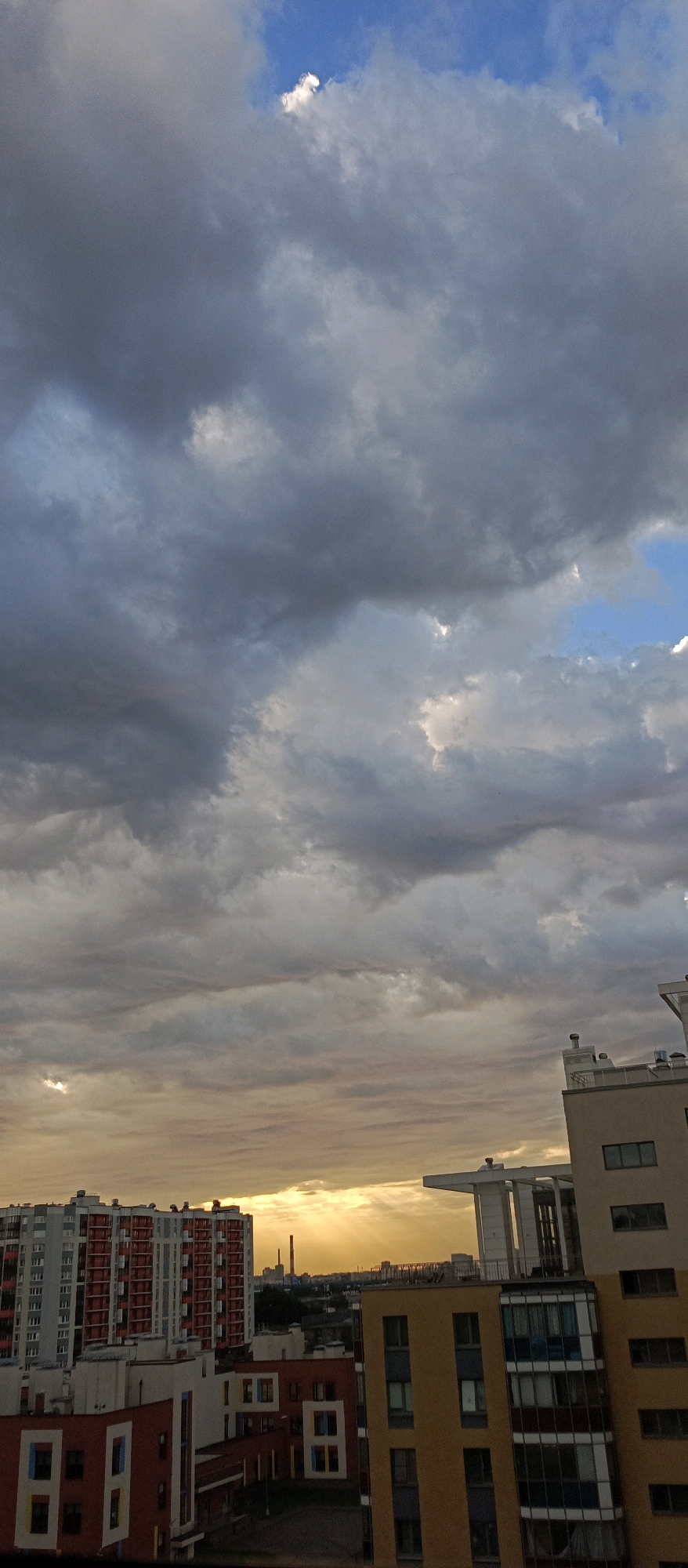 An aerial, eye-level, vertical medium shot captures a city skyline under a dramatic sky. Dark, heavy clouds dominate the upper portion of the frame, with patches of blue sky visible and rays of sunlight breaking through. In the foreground, modern apartment buildings of varying heights and colors are visible, including a tall building with red and white accents and other buildings in yellow and white. A chimney stack is visible in the distance, beyond the buildings, with industrial structures in the background. The lighting suggests either dawn or dusk, with the sun's rays creating a dramatic effect. There are no people or animals visible in the frame. The scene is set in an urban environment.