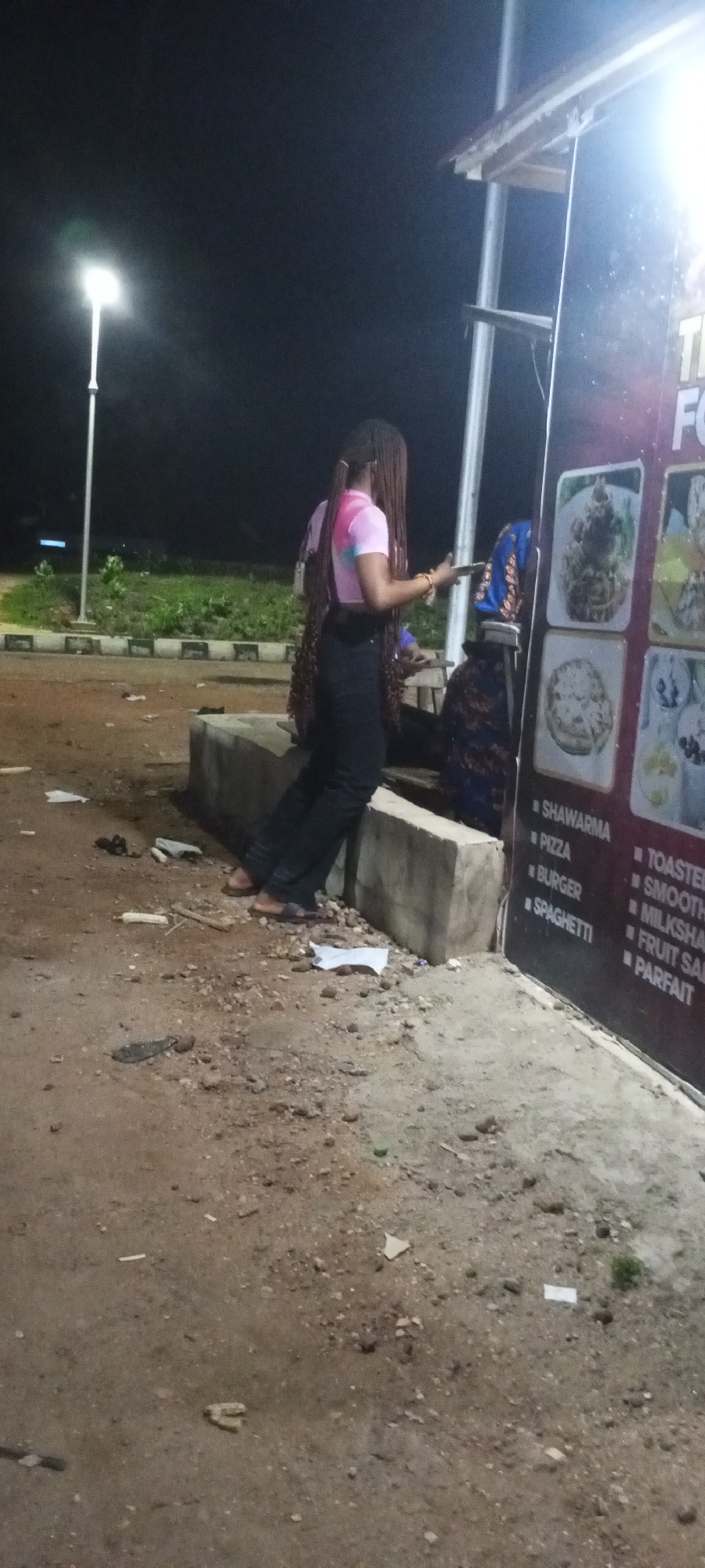 A young Black woman with braided hair, wearing a pink and white tie-dye crop top and black pants, stands at night outdoors in Adankolo, Nigeria. She is holding a mobile phone in her right hand. Behind her, another person wearing a blue and purple patterned garment is partially visible. They are standing next to a concrete structure. To the right, a large black sign displays images of food items and text. The ground is uneven and covered with dirt, rocks, and debris, including pieces of white paper and a discarded ear of corn. In the background, a street lamp illuminates the scene. The overall impression is of an informal outdoor setting, possibly a food stall or roadside eatery at night.