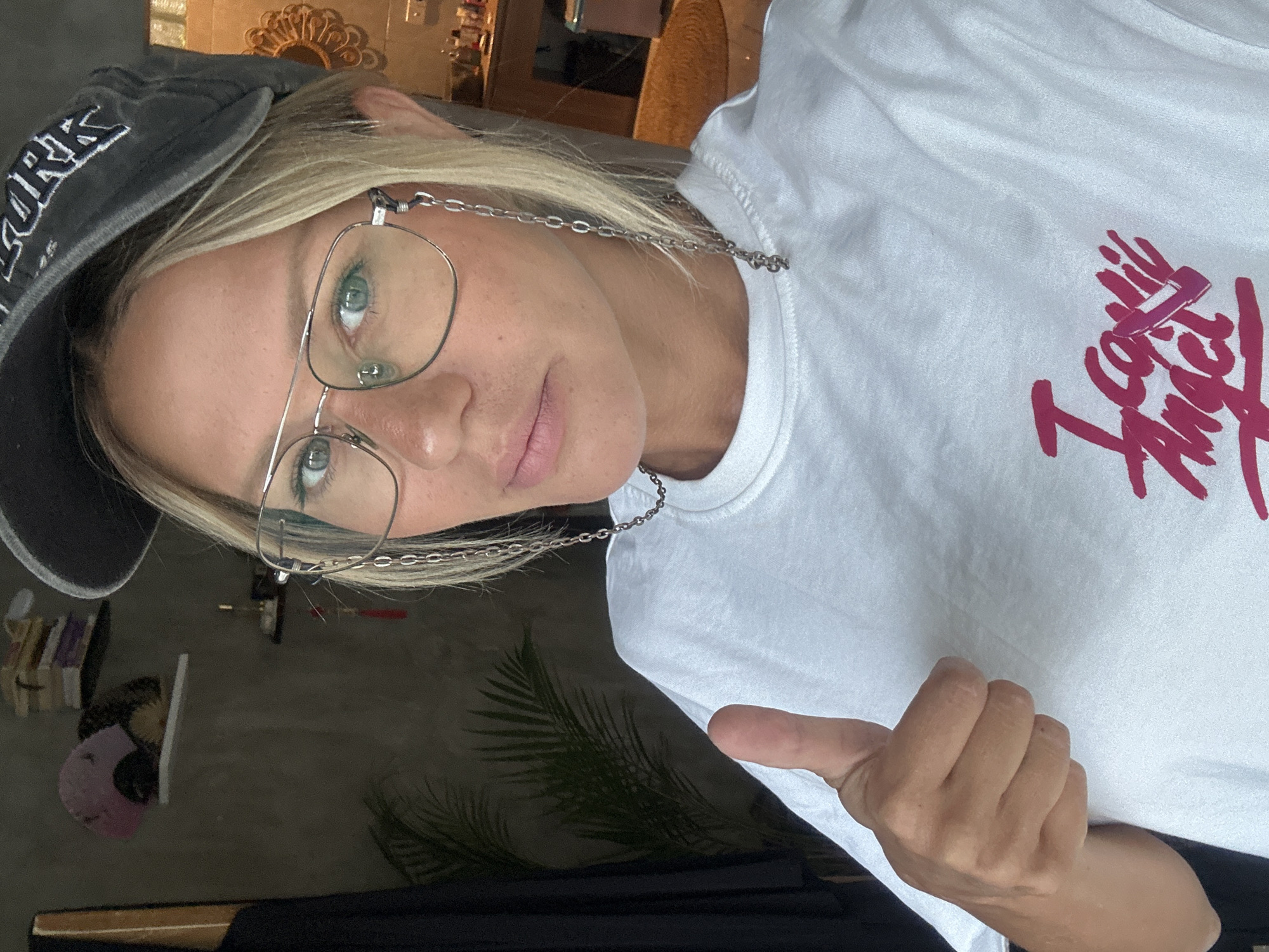 A woman with blonde hair, wearing a grey baseball cap with "New York" printed on it, eyeglasses, and a white t-shirt with "I call Angel" printed in pink, is giving a thumbs up. The background includes a palm leaf and a partial view of a wooden dresser. It cannot be verified from the image that the location is North Kuta, Indonesia.