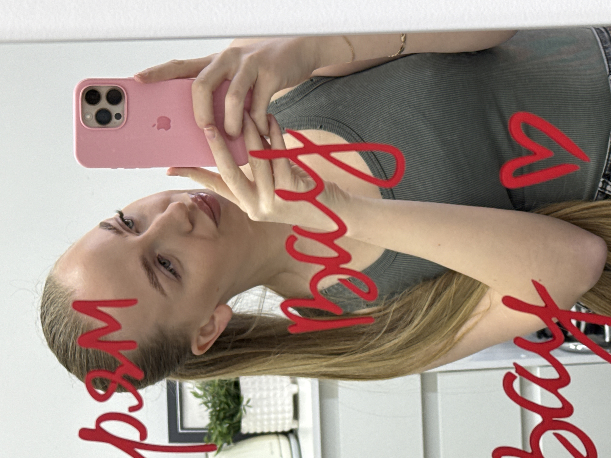 An adult female subject, appearing to be in her 20s or 30s, is depicted taking a mirror selfie in an indoor setting within the city of Petropavlovsk-Kamchatsky, Russia. The subject has long, light brown hair styled in a ponytail and is wearing a dark olive green ribbed tank top. She holds a pink-cased iPhone (featuring a triple-lens camera module) with both hands, positioned in front of her torso. Her gaze is directed towards the phone's screen or slightly to the side. A thin gold-colored chain bracelet is visible on her left wrist.
The mirror's surface features handwritten text and a symbol in red. From top to bottom, the visible text reads "pяM", followed by "bay", and then another "bay", with a heart symbol below the second "bay". This text, as observed in the reflection, implies that the original writing on the mirror, intended to be read correctly by the writer, would be "MяP" (Мир - meaning "World" or "Peace" in Russian) and two instances of a word that appears as "Bay" when viewed directly.
The background, visible as a reflection in the mirror, includes a white wall and a white shelving unit on the left. On the shelf, a white textured pot holds a small green plant, with a partial black picture frame visible behind it. Further to the left, a portion of a white kitchen appliance, possibly a kettle, is present. The overall lighting suggests an interior room environment.