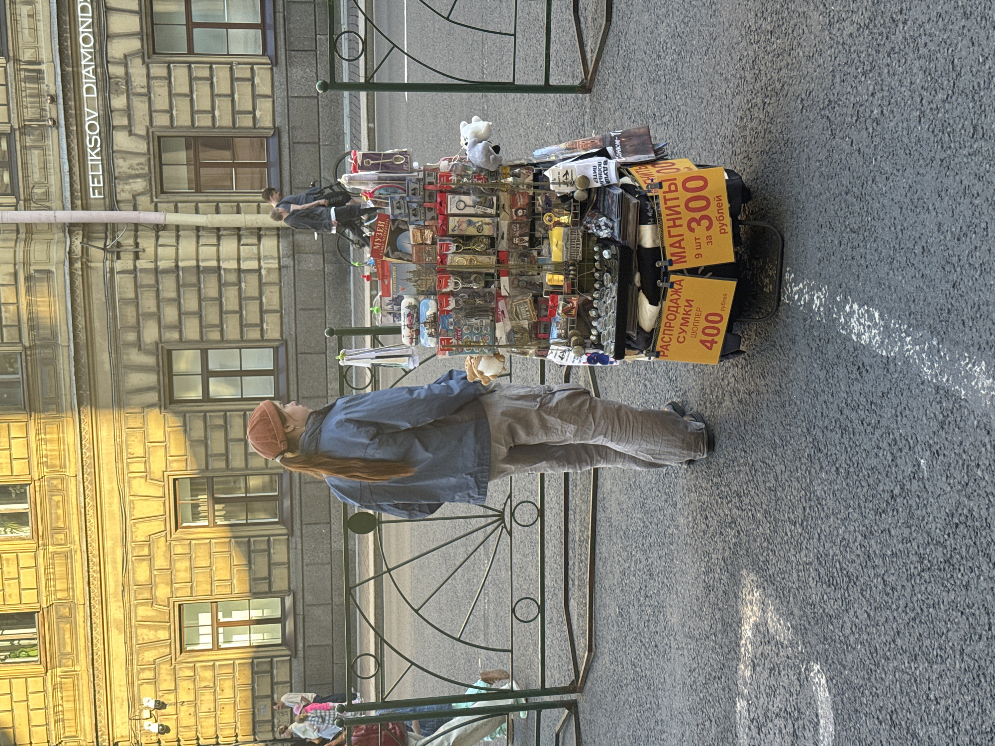 A young woman with reddish-brown hair in a ponytail, wearing a blue jacket, beige cargo pants, and a burnt orange cap, stands on a sidewalk in Saint Petersburg, Russia. She is facing away from the camera, looking towards a street where a vendor's cart is set up. The cart is laden with souvenirs, including magnets, keychains, and miniature replicas of landmarks. Two yellow signs are attached to the cart, one reading "РАСПРОДАЖА СУМКИ ШОППЕР 400 рублей" (Sale of tote bags, 400 rubles) and the other "МАГНИТЫ 9 шт за 300 рублей" (Magnets, 9 pieces for 300 rubles). In the background, a building with a yellow, textured facade and large windows stands tall. Several people are visible walking on the sidewalk and street. The scene is illuminated by sunlight, casting shadows on the building and sidewalk.