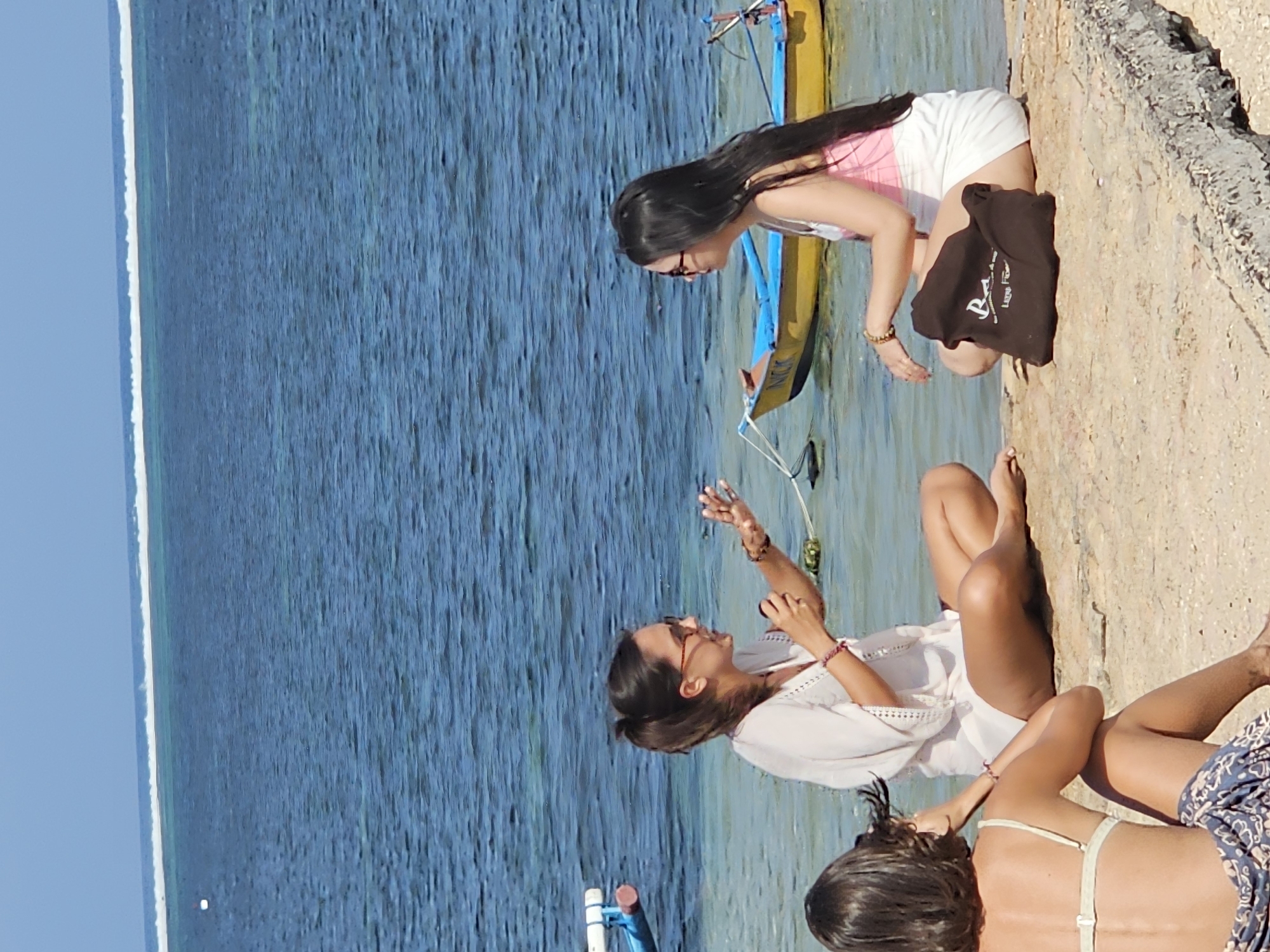 Three individuals are seated on a concrete ledge near the ocean. The individual on the far left, seen from behind, wears a light-colored bikini top and a patterned sarong. The central figure, a woman with dark hair pulled back in a ponytail and wearing glasses, is dressed in a sheer white tunic and poses with her hands outstretched as if speaking. The third individual, to the right, has long dark hair, wears glasses, and is dressed in a pink tank top and white shorts. A dark brown tote bag rests on the ledge beside her. Two boats are visible in the shallow, blue water: a smaller, blue and yellow vessel and a portion of a white and blue boat. The background consists of the ocean and a clear sky. The scene is located on the beach in Denpasar, Indonesia.