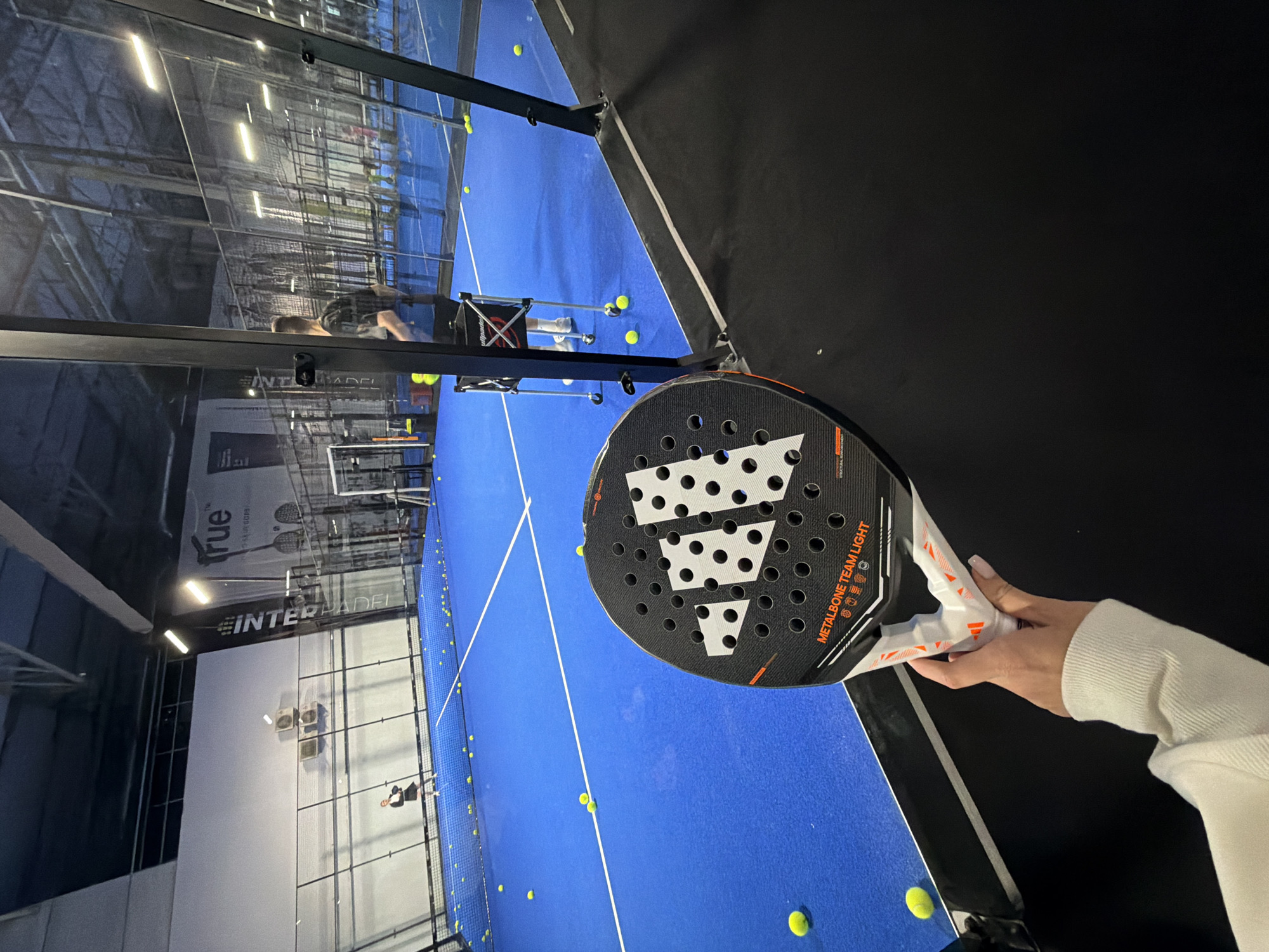 The image captures a view from within an indoor padel court, possibly in a city of Unknown, Unknown. The foreground features a hand holding an Adidas Metalbone Team Light padel racket, its design prominently displayed. The court itself is a vibrant blue, with scattered yellow padel balls adding to the active scene. In the background, a person is seated on a chair, engaged in observing the game, or potentially resting. The court is enclosed with clear glass barriers, which reflect the lighting and framework of the facility. Visible through the glass walls are advertisements or branding, including "INTERPADEL," suggesting a commercial padel facility. The contrast between the well-lit court and darker areas enhances the dynamic composition of the image.