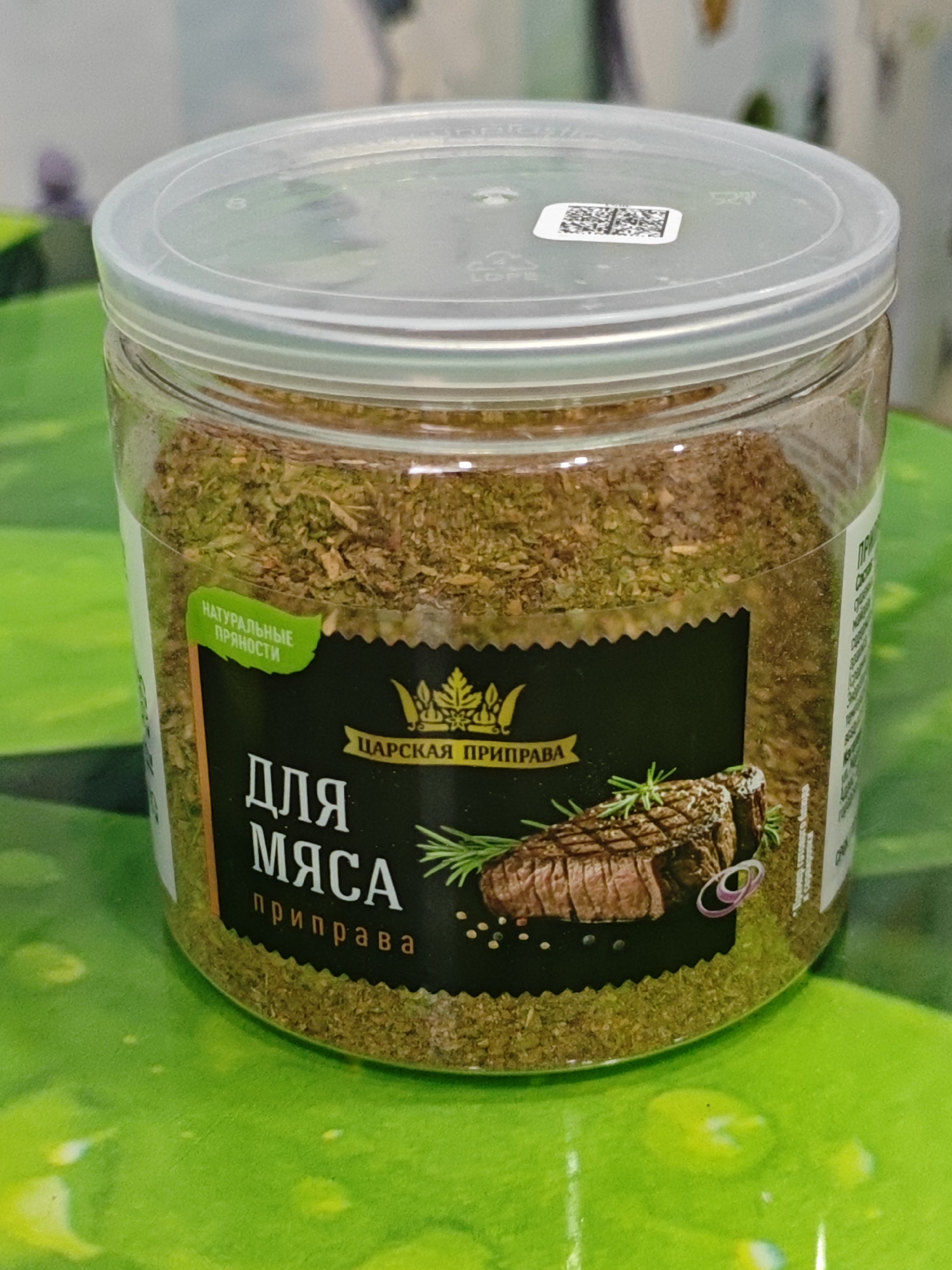 The image shows a clear plastic jar containing a dry spice mix. The jar has a transparent lid with a QR code sticker. A black label is adhered to the front of the jar. The label has text in Russian and features an illustration of a steak with rosemary and peppercorns. The text on the label reads "ЦАРСКАЯ ПРИПРАВА" (Tsarskaya Priprava - Royal Seasoning) and "ДЛЯ МЯСА" (Dlya Myasa - For Meat), with "приправа" (priprava - seasoning) written below. A green sticker on the left side of the jar says "НАТУРАЛЬНЫЕ ПРЯНОСТИ" (Natural Spices). The jar is placed on a bright green, reflective surface, possibly a table. The background is blurred, with hints of white and pastel colors. There is no indication of the city of Polyarny, Russia, within the image.
