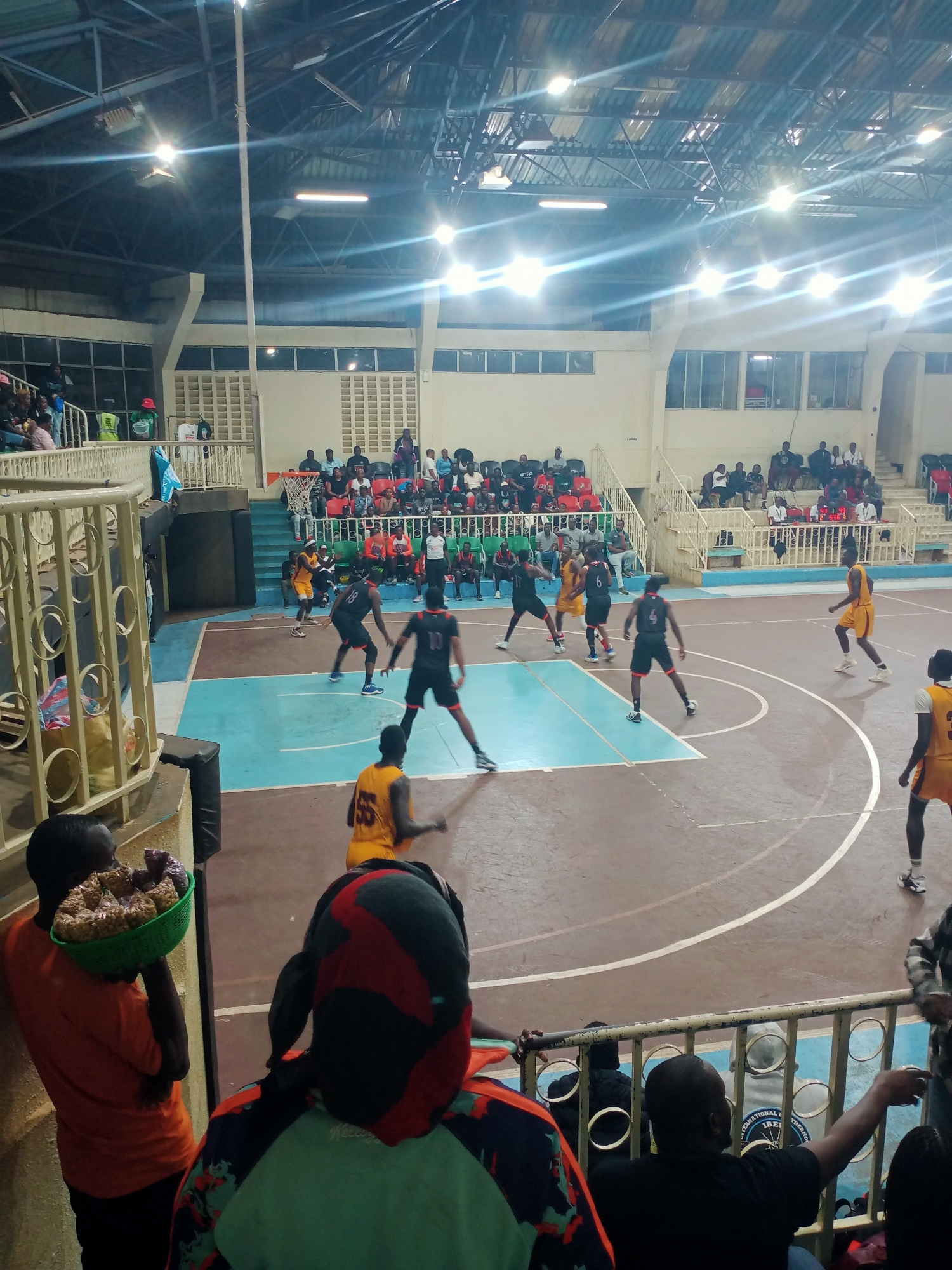 The image depicts a basketball game in progress at an indoor court. Players in black uniforms with red accents are facing off against players in yellow uniforms. Spectators are seated in tiered stands around the court. A man in an orange shirt stands in the foreground carrying a green basket filled with what appears to be food items. People in the foreground are watching the game. The venue has a high ceiling with artificial lighting and a visible metal framework. The location is likely in Mugumo-ini ward, Kenya.