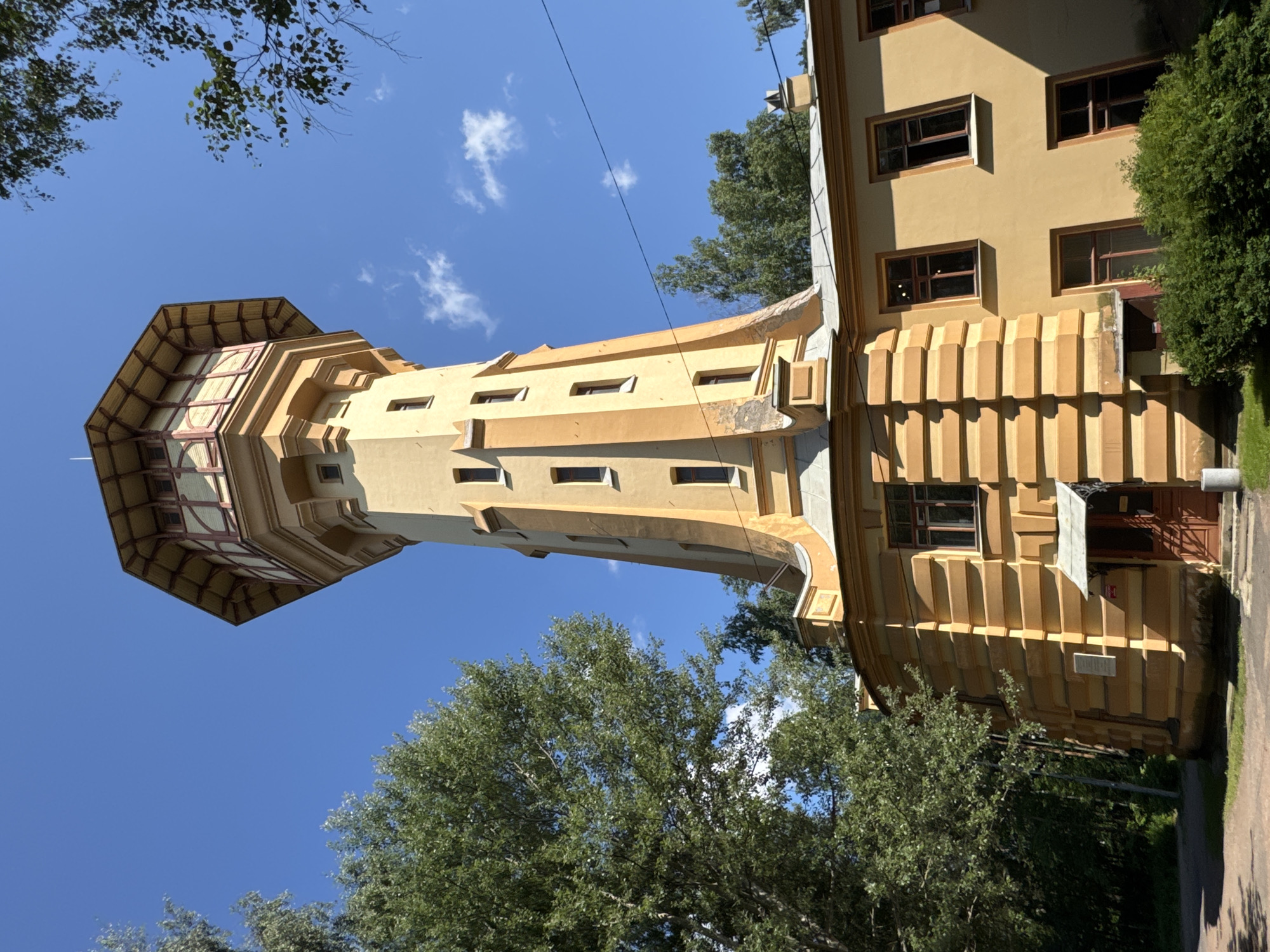 The image shows the Vitebsky railway station water tower in Saint Petersburg, Russia, on a bright, sunny day with a clear blue sky and scattered white clouds. The tower, a prominent ochre-colored structure with ornate detailing, stands tall against the sky. It features a tapered octagonal top section with a decorative roof and numerous windows. The lower part of the tower is attached to a yellow, rectangular building with a similar architectural style, characterized by horizontal banded decorative elements and multiple windows. Green trees and foliage surround the building, partially obscuring it on the left and right sides. The ground in front of the building appears to be a paved path or courtyard, with some grass and bushes. The scene is devoid of any people or visible activity, presenting a static architectural view.
