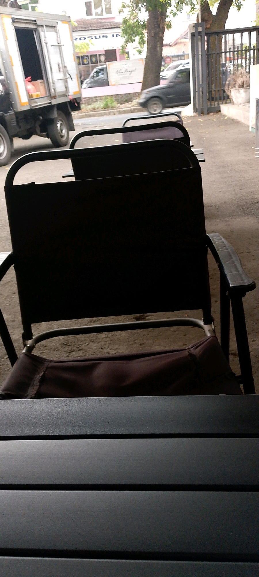 In Bandung City, Indonesia, an outdoor scene unfolds near a roadside establishment, possibly a cafe or restaurant. The foreground is dominated by the back of a dark-colored, foldable chair, its fabric seat a deep brown and its metal frame slightly visible. A dark, slatted table occupies the very bottom of the frame, suggesting a seating area.
Beyond the chairs, a road is visible, with a light-colored truck on the left, its rear doors open and showing some contents. Further down the road, a dark-colored sedan is in motion. The background features buildings with signs, including one that appears to read "ROSUS IN". A mature tree with a thick trunk stands prominently to the right, with foliage that creates shadows. A dark metal gate and a potted plant are also present. The ground is a mix of asphalt and dirt. The overall lighting suggests an overcast day.