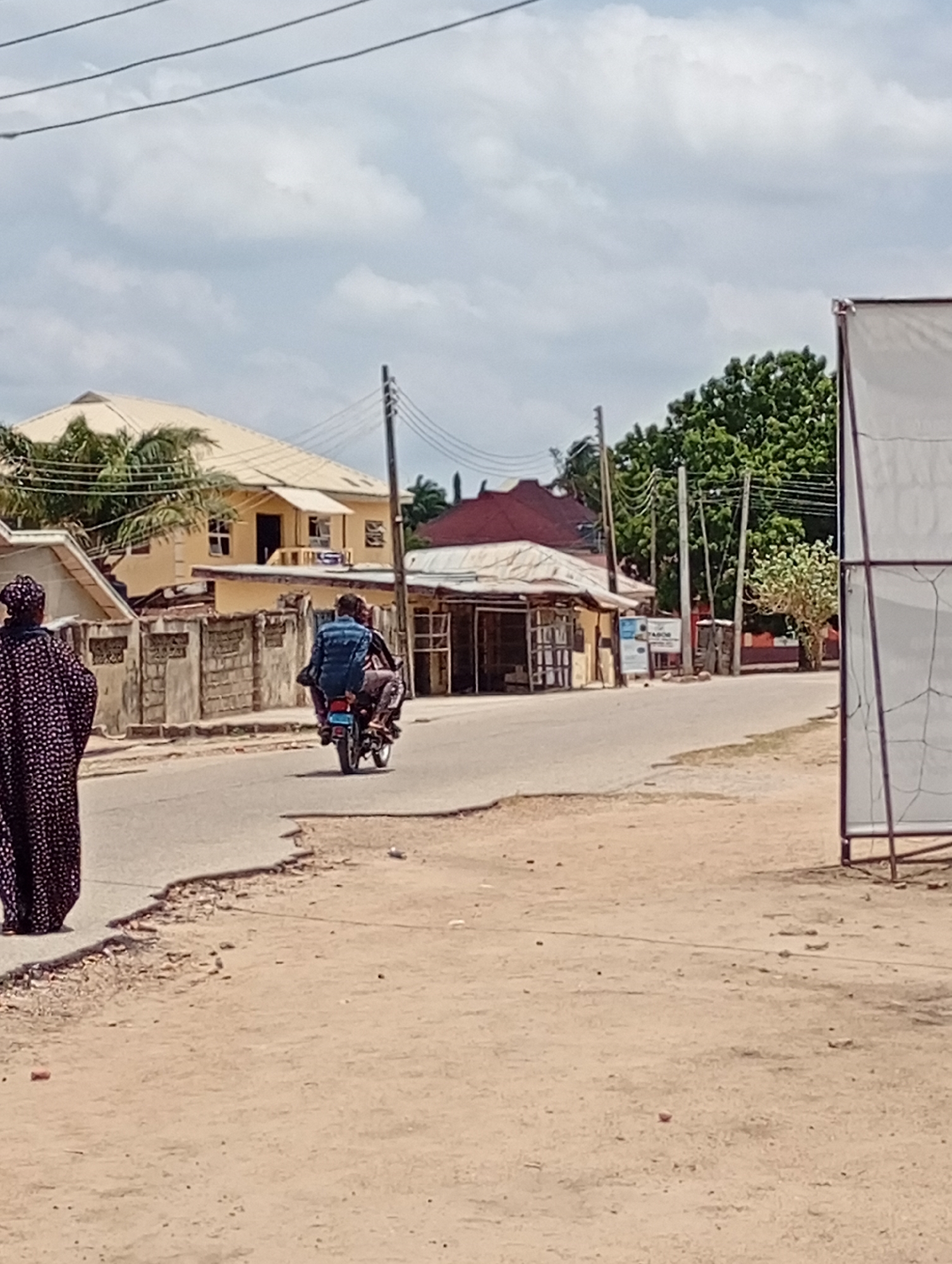 A medium-angle daytime shot depicts a street scene in Lokoja, Nigeria. The foreground shows a sandy, uneven ground with sparse debris. To the left, a woman wearing a long, dark patterned robe walks along the edge of the road.
In the middle ground, a motorcycle with two occupants is moving away from the camera on the paved road. The rider is wearing a dark shirt, while the passenger behind him is wearing a blue shirt and carrying a backpack.
Buildings line both sides of the street. On the left, there's a yellow two-story building with a light-colored roof behind a light colored concrete fence. Next to it is a smaller building with a corrugated red roof and open storefront. On the right, there are trees and buildings further down the road. Utility poles and wires are visible overhead.
A white structure with a metal frame is present in the right foreground. The sky is cloudy.