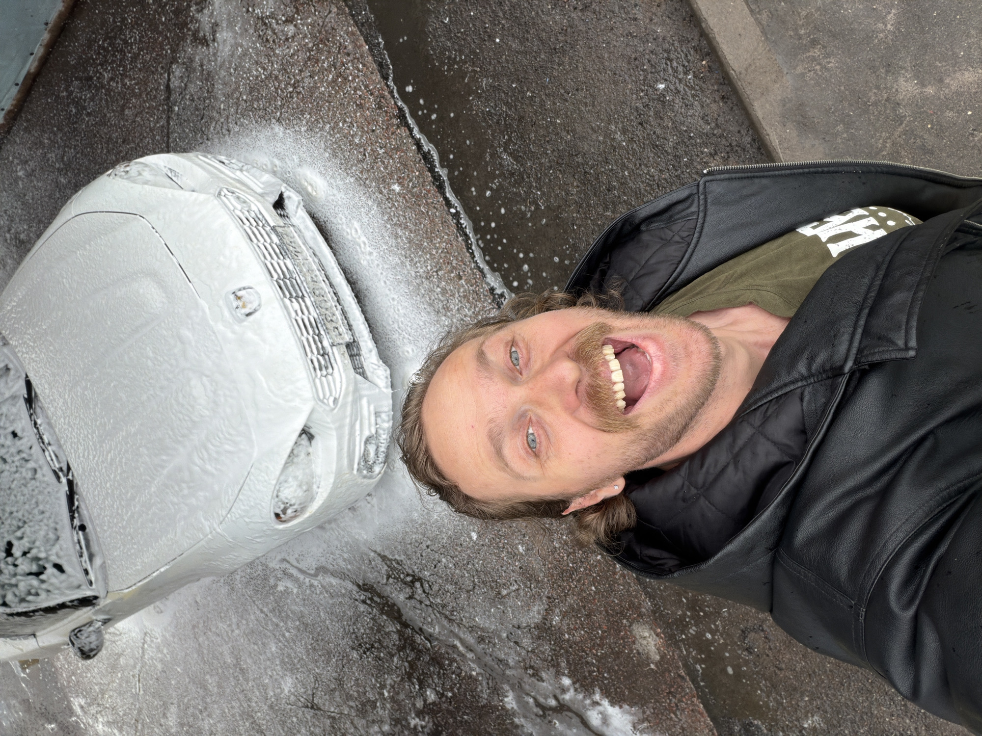 The image depicts a man taking a selfie next to a car covered in foam. The man has light skin, blue eyes, and a mustache. He is wearing a black leather jacket over a green shirt with white lettering. He is looking at the camera with his mouth open in a wide grin.
To the left of the man is a white car entirely covered in white foam. The car appears to be parked on the side of a street.
The ground is wet and there is runoff from the foam, suggesting the car was recently washed. The background is a concrete surface, possibly a sidewalk or the edge of a road.
Given the architectural style of the car, it is identifiable as a Fiat 500, a modern remake of the classic version. This can be used as an identifying detail to connect this photo to the city of St. Petersburg, Russia.