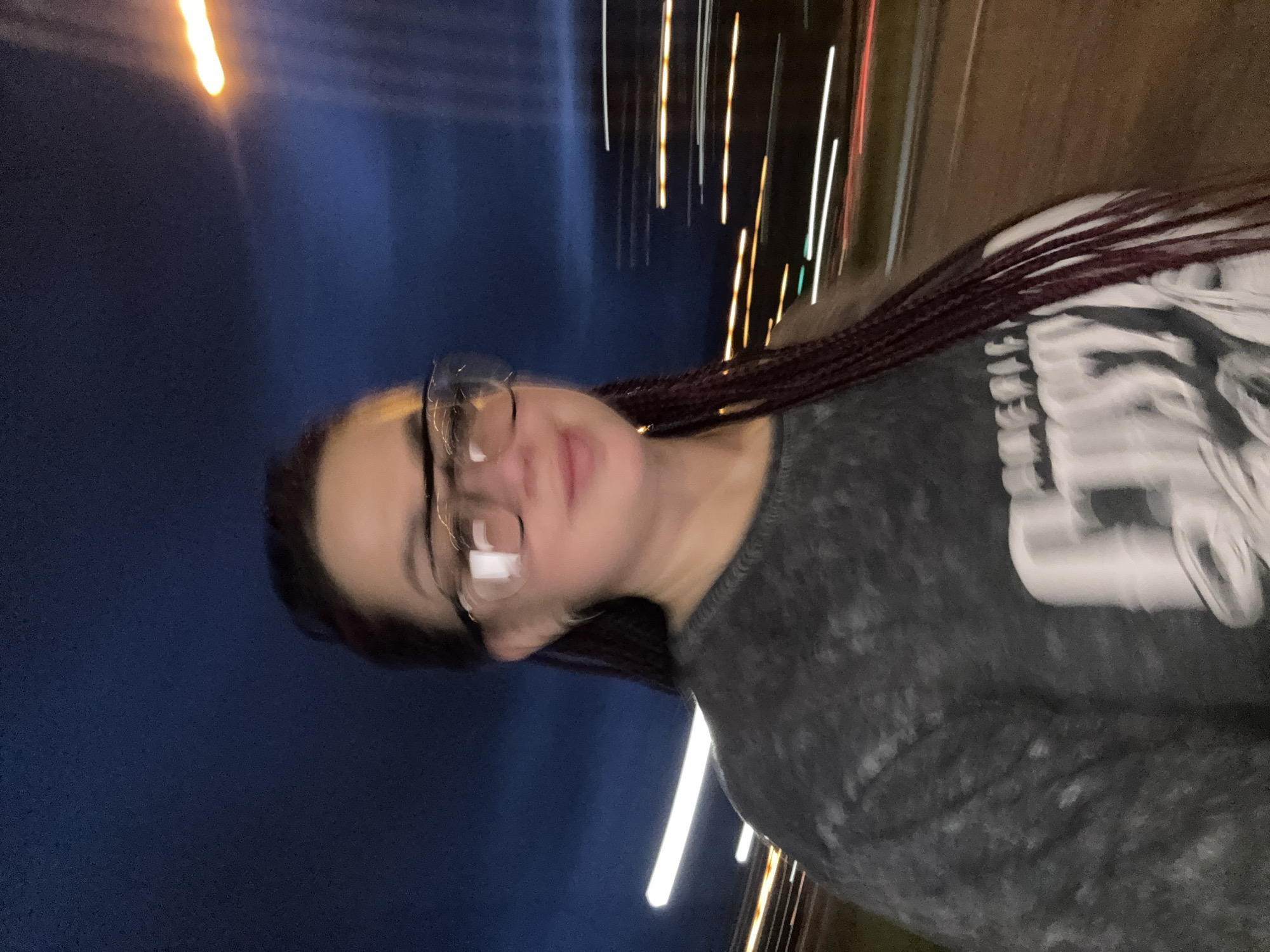 The image shows a person wearing glasses and a dark gray t-shirt with a graphic design on the front. The person has dark hair styled in braids. The background suggests an outdoor urban environment at night, with streaks of light indicating movement, possibly from vehicles on a street or highway. The sky is dark blue, and there are streetlights or vehicle lights visible as blurred lines of white and orange. The image appears to have been taken while the person was in motion. Without additional context, it is impossible to determine the exact location within Tolyatti, Russia.