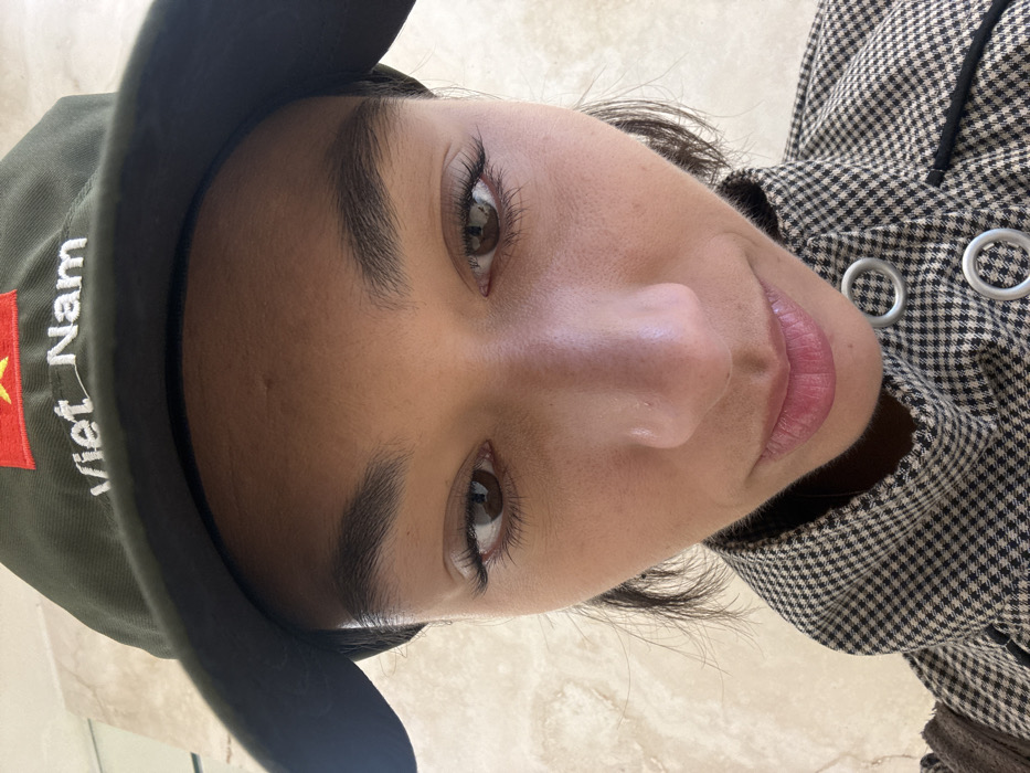 A close-up, downward-angled photographic portrait shows an individual from the eyes to the upper chest. The person has a medium-dark skin tone, dark brown eyes with visible eyelashes, and a small silver stud piercing on the left nostril. They wear a dark green baseball cap featuring the red and yellow star flag of Vietnam, with "Viet Nam" embroidered in white text beneath it. Dark hair is visible extending from under the cap. The individual is dressed in a black and white (or dark brown and cream) small checkered pattern jacket or coat, which includes prominent silver-colored circular grommets and black piping on the front. The background consists of a uniformly light-colored, slightly textured wall, which offers no distinct architectural or environmental cues. The specific location within Paris, France, cannot be determined from the visual information provided.