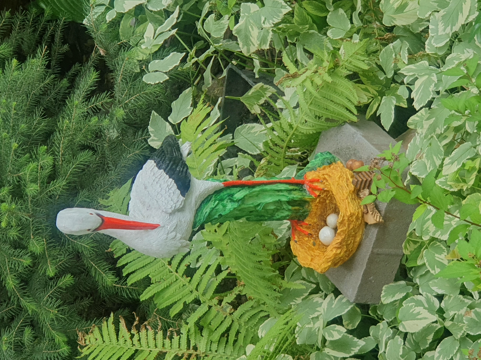 An ornamental garden sculpture of a stork stands prominently in a lush green garden. The sculpture depicts a white stork with black wingtips and a red beak and legs, perched on a green, tree-like base. Below the stork, a yellow nest contains two white eggs. The sculpture is situated on a grey concrete block amidst a variety of green foliage, including ferns and other leafy plants. The background is filled with dense evergreen branches. The scene is a close-up shot, focusing on the stork and its immediate surroundings, with no people visible. The location is a garden in the city of Almaty, Kazakhstan.
