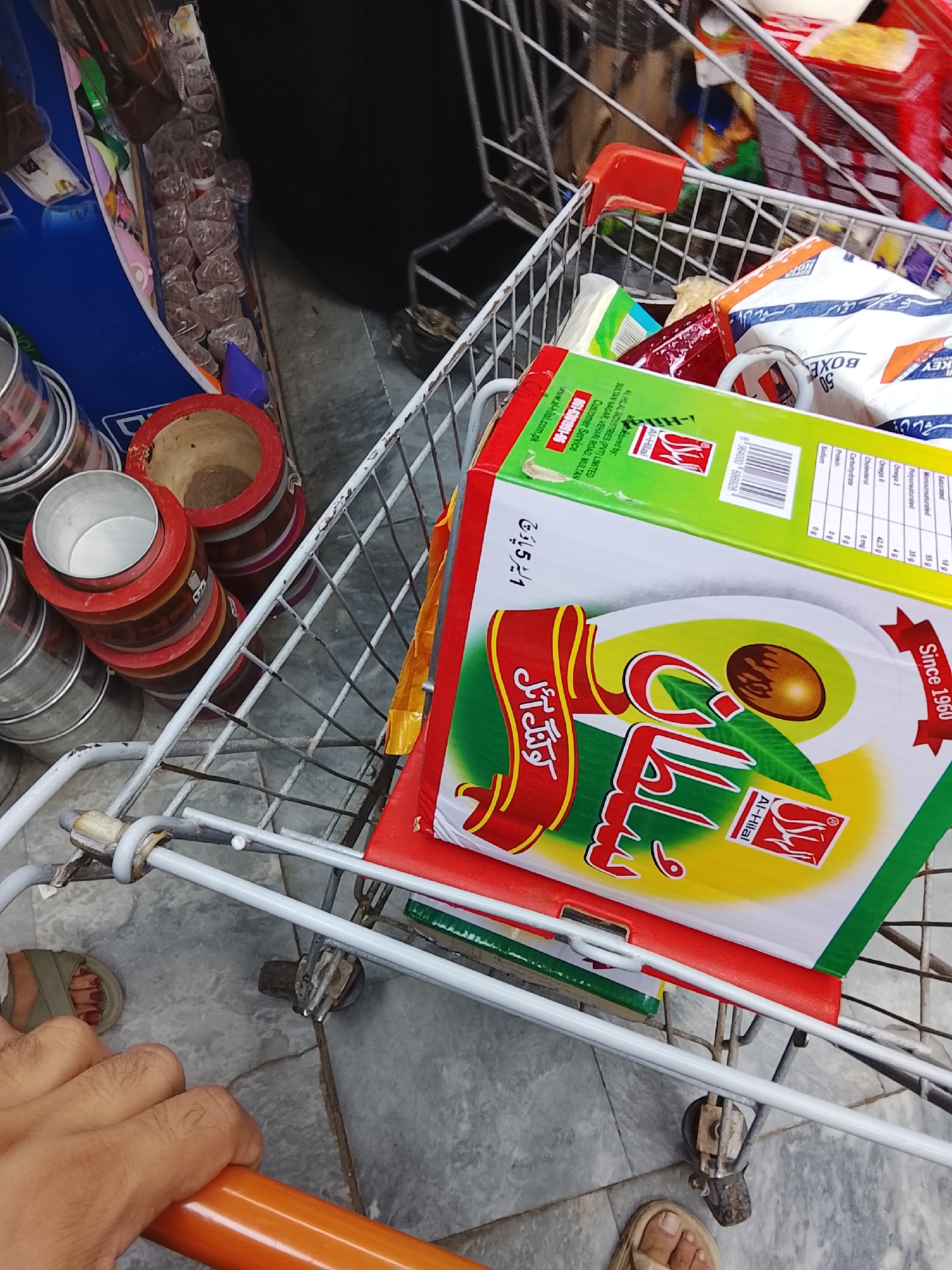 A close-up, overhead shot depicts a shopping cart mostly filled with groceries. At the bottom of the cart is a rectangular box of Al-Hilal Sultan cooking oil, a Pakistani brand. The box is red, white, green, and yellow, displaying the brand name and an avocado image. Additional grocery items such as snacks and packages are visible but not clearly identifiable. Adjacent to the cart on the left, are stacked metal containers and red-banded wooden planters. In the bottom left and right corners of the image, portions of two feet (sandaled) and a hand holding the shopping cart handle, are visible. The setting is on a concrete surface, possibly within a grocery store. This is not directly visible, the scene is close up to the cart. There is no visible direct evidence of the location being Gulgasht Colony, Pakistan.