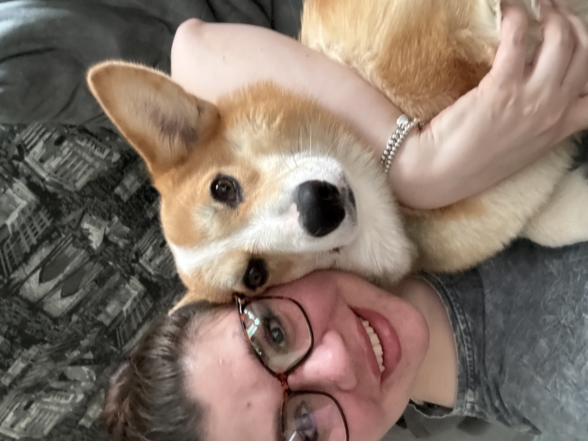 A woman with brown hair pulled back and wearing glasses is lying on a bed with a gray patterned blanket. She is smiling and holding a corgi dog close to her face. The dog has brown and white fur and is looking at the camera. The woman is wearing a gray shirt and a silver bracelet. There is no indication of this image being taken in Votkinsk, Russia.
