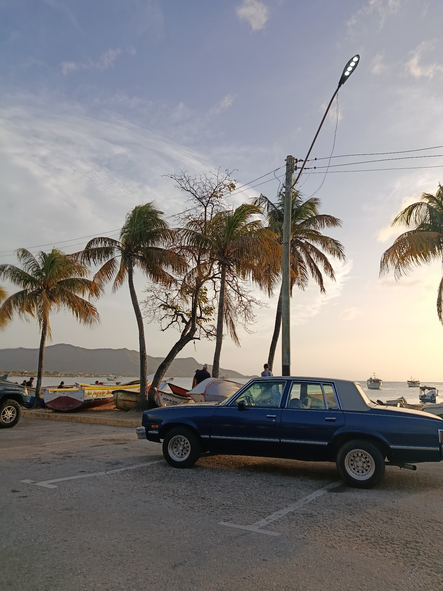 The image depicts a street scene in Juan Griego, Venezuela, likely during the late afternoon or early evening. In the foreground, a dark blue four-door sedan is parked on an asphalt surface marked with white parking lines. The car has chrome trim and whitewall tires. To the left, the front tire and part of a dark-colored SUV are visible.
Behind the vehicles, a waterfront area is visible with several small fishing boats pulled up onto the shore. Palm trees line the area, some with fronds blowing in the wind. In the background, a body of water, possibly the sea, is dotted with several boats, and a mountainous coastline can be seen in the distance. The sky is a pale blue with scattered clouds, and the lighting suggests either sunrise or sunset. A street light with multiple LED bulbs stands on a pole near the palm trees. Several people are scattered around the boats, some appearing to be interacting with them or sitting nearby. One person stands near the parked sedan, looking out towards the water.