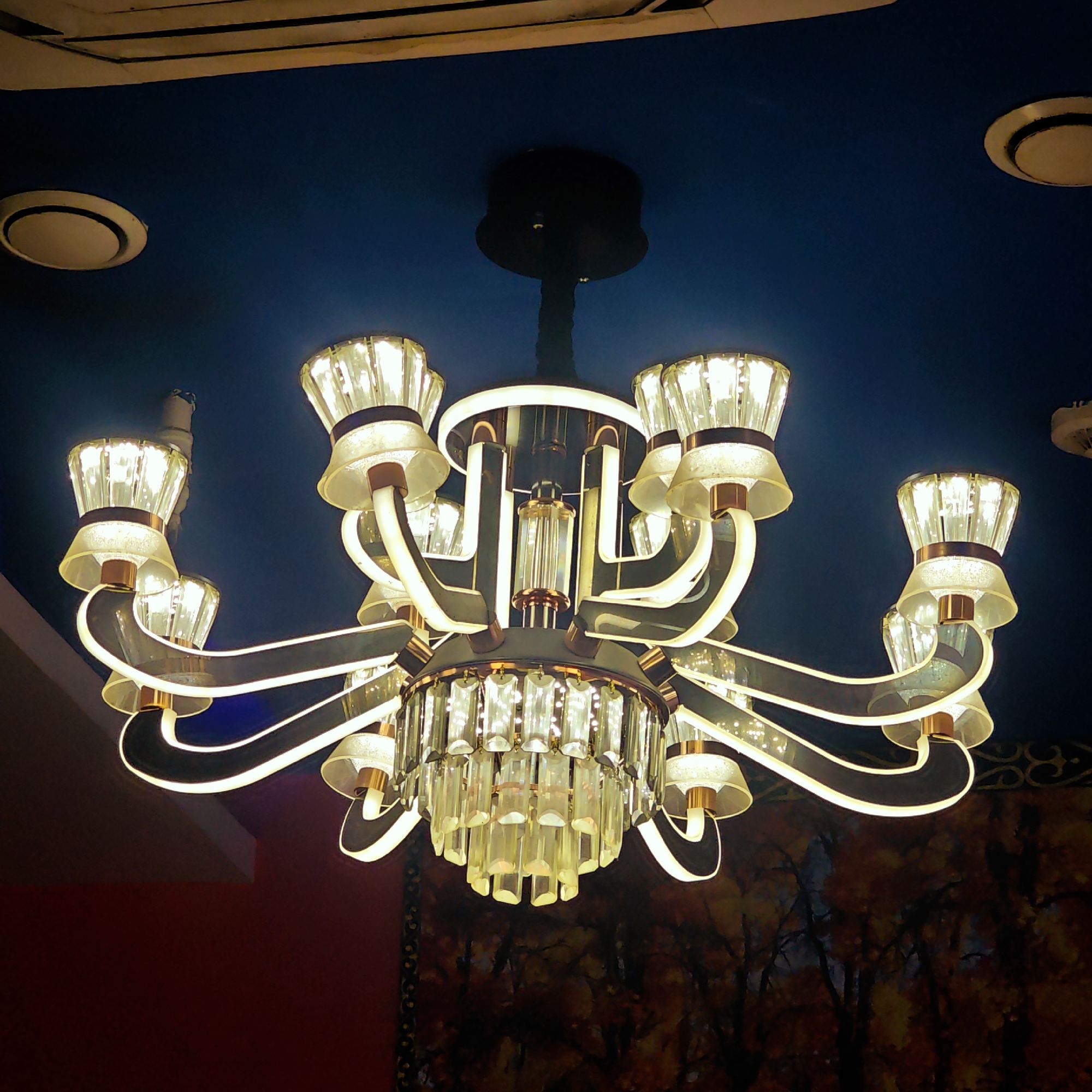 A chandelier with multiple glass shades and crystal pendants hangs from a blue ceiling in a room, likely in Yuzhong District, China. The chandelier has curving, illuminated arms and a central structure surrounded by hanging crystals. Two circular recessed lights are also visible in the ceiling. Below the chandelier, the bottom part of a wall has a dark red color while the remaining area displays a mural resembling a forest or trees in autumn.