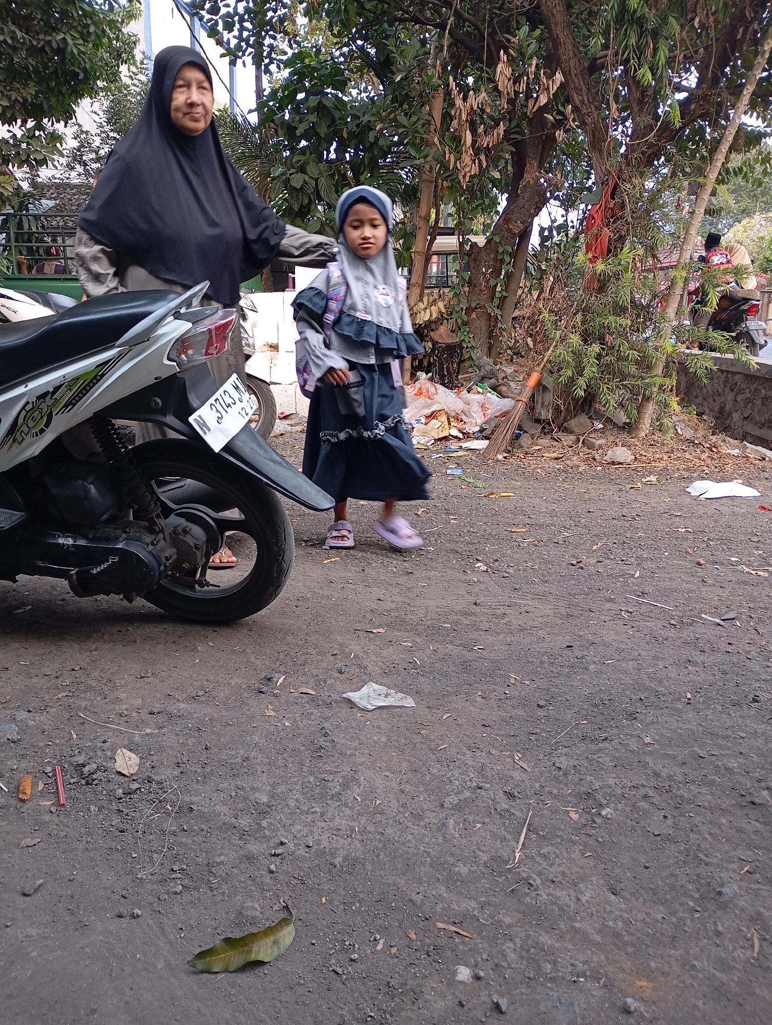 In Ketegan, Indonesia, a woman wearing a black hijab stands next to a young girl in a gray hijab and dress, who carries a backpack. They are positioned in front of a white motorcycle, with its license plate reading "N 3743 MJ". The background features trees, foliage, and a stone wall. The ground is dirt and gravel, with scattered debris including leaves, plastic, and a broom.