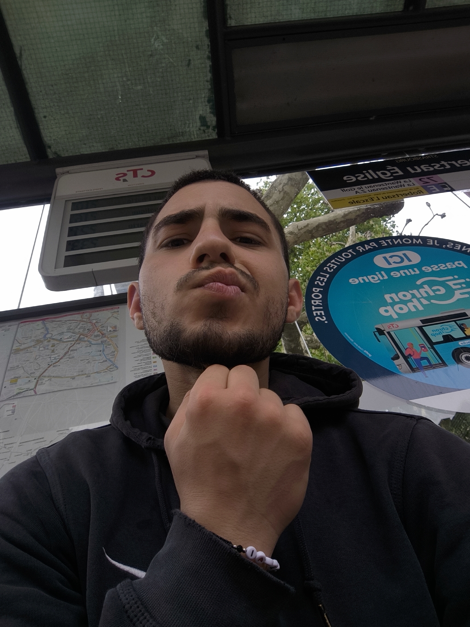 A man is taking a selfie at a Compagnie des Transports Strasbourgeois (CTS) bus stop in Strasbourg, France. The bus stop displays CTS logos, a map of the public transportation network, and bus route information. The man is wearing a black hoodie with a white Nike swoosh, and a beaded bracelet on his left wrist. He has a beard and his hand is touching his chin.