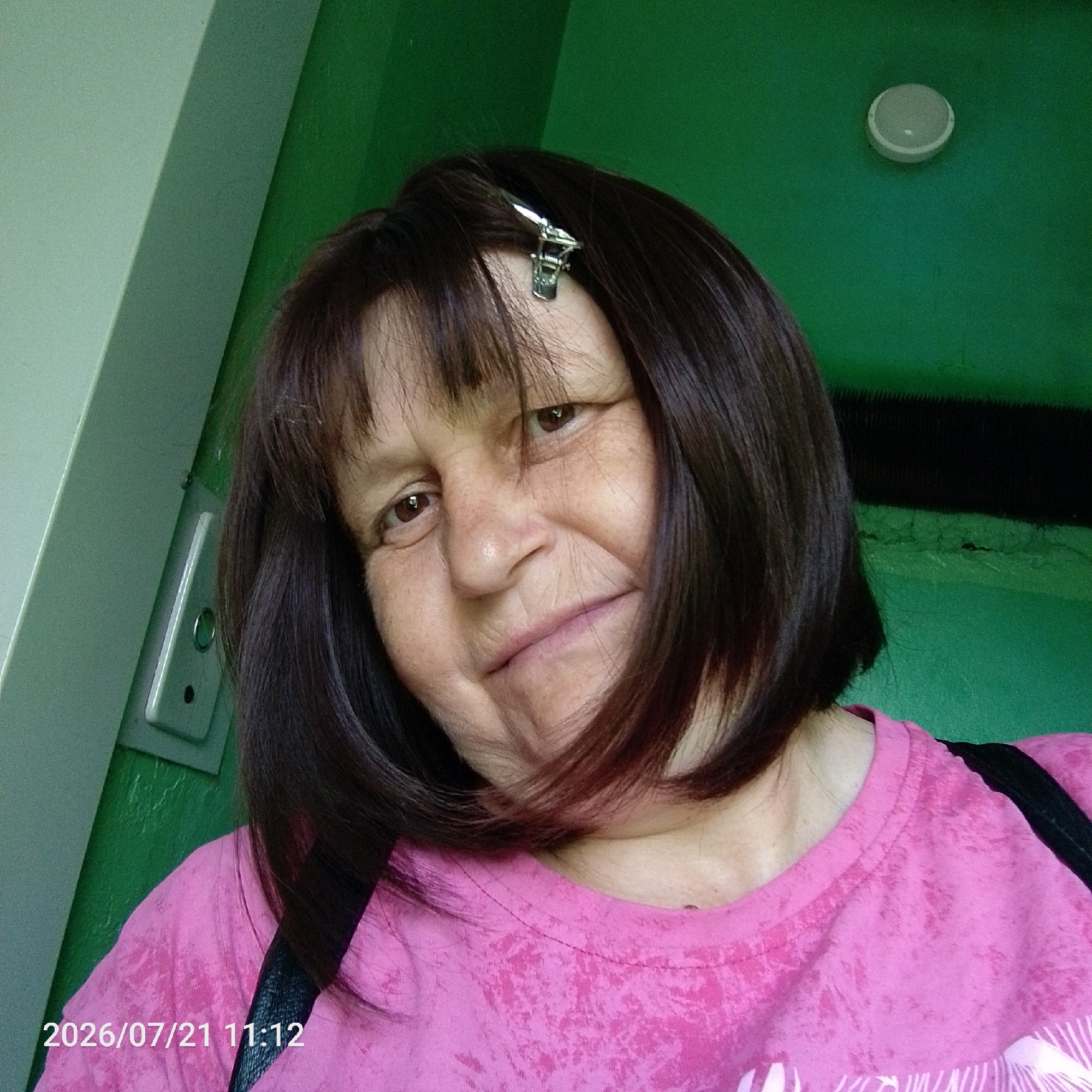 A medium shot image shows a woman with brown hair and bangs, a hair clip in her bangs, and a pink t-shirt. The background is a green wall with a light fixture. The timestamp on the image reads 2026/07/21 11:12. The woman's face is partially obscured by her hair. She is smiling.