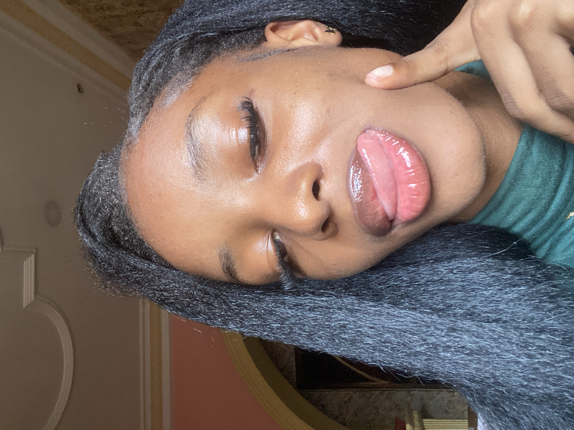 A young Black female subject is depicted in a close-up, chest-up selfie-style photograph, taken in an indoor setting located in Jos, Nigeria. The subject has a mid-to-deep skin tone and long, dark, braided or textured hair styled to fall over her left shoulder (viewer's right). Her eyes are partially closed or downcast, adorned with long, dark eyelashes, likely extensions. Her lips are full, coated in a glossy pink product, and her tongue is extended upwards, appearing to lick her upper lip. The index finger of her right hand (viewer's left) is gently pressed against her right cheek. She is wearing a dark green, ribbed turtleneck garment. The background reveals architectural details of the interior space: to the subject's left, a light peach-colored wall is visible, with a portion of a golden, ornate archway curving below it. The ceiling above is light-colored and features prominent white decorative moldings, including a semicircular design and parallel linear strips. To the subject's right, a section of a textured or patterned wall, possibly wallpaper, in varying shades of brown and gold, is visible.