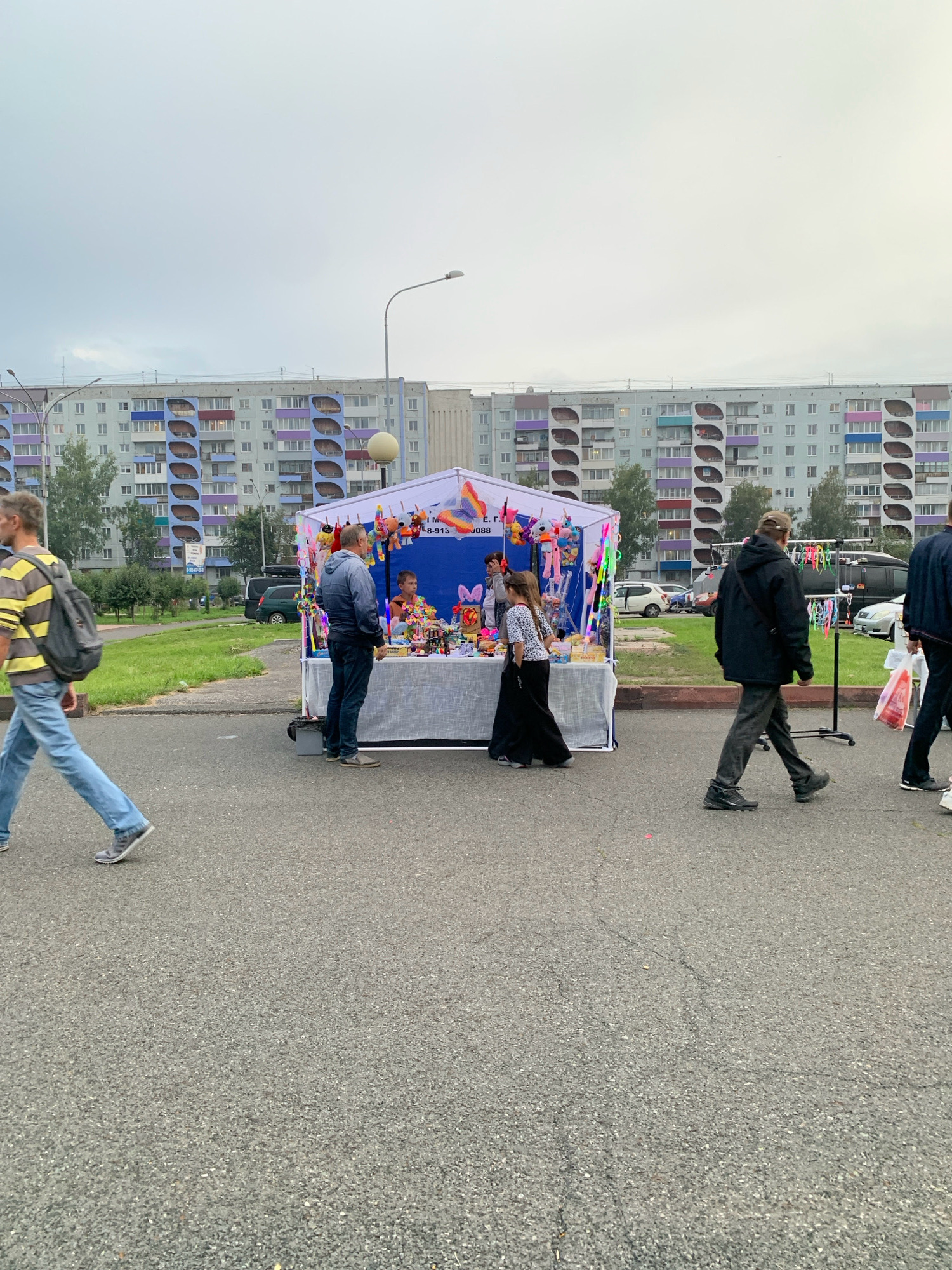 The image depicts an outdoor market stall in Prokopyevsk, Russia, in front of a tall, multi-story apartment building with a distinctive blue and purple facade. The sky is overcast. The stall is a temporary structure with a white tent and a blue backdrop. It is laden with various small items, including plush toys, novelty items, and possibly candy. Several people are visible around the stall. A man in a striped shirt and backpack walks past in the foreground, to the left. A man in a blue jacket stands at the stall, interacting with a younger boy who is also at the stall. A woman in a black skirt and white patterned top is also at the stall, appearing to be a vendor. To the right, two men are walking away from the stall. One is wearing a black jacket and cap, and the other is carrying a white shopping bag. In the background, there are several parked cars and a green grassy area. The overall scene suggests a local community event or market day.